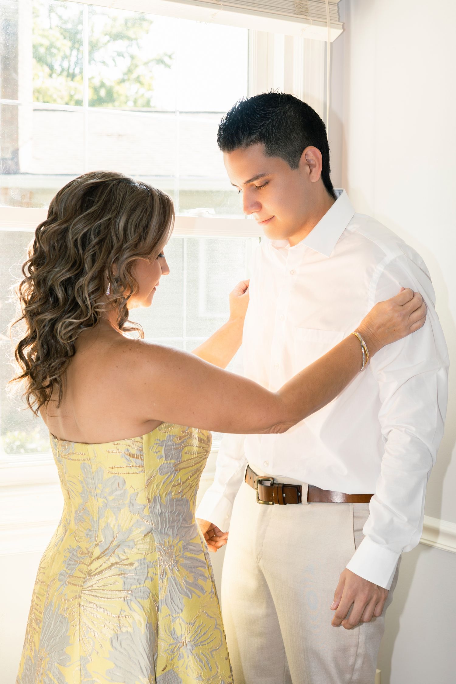 A couple shares an intimate moment by a window, with her wearing a yellow floral dress and him in a white shirt.