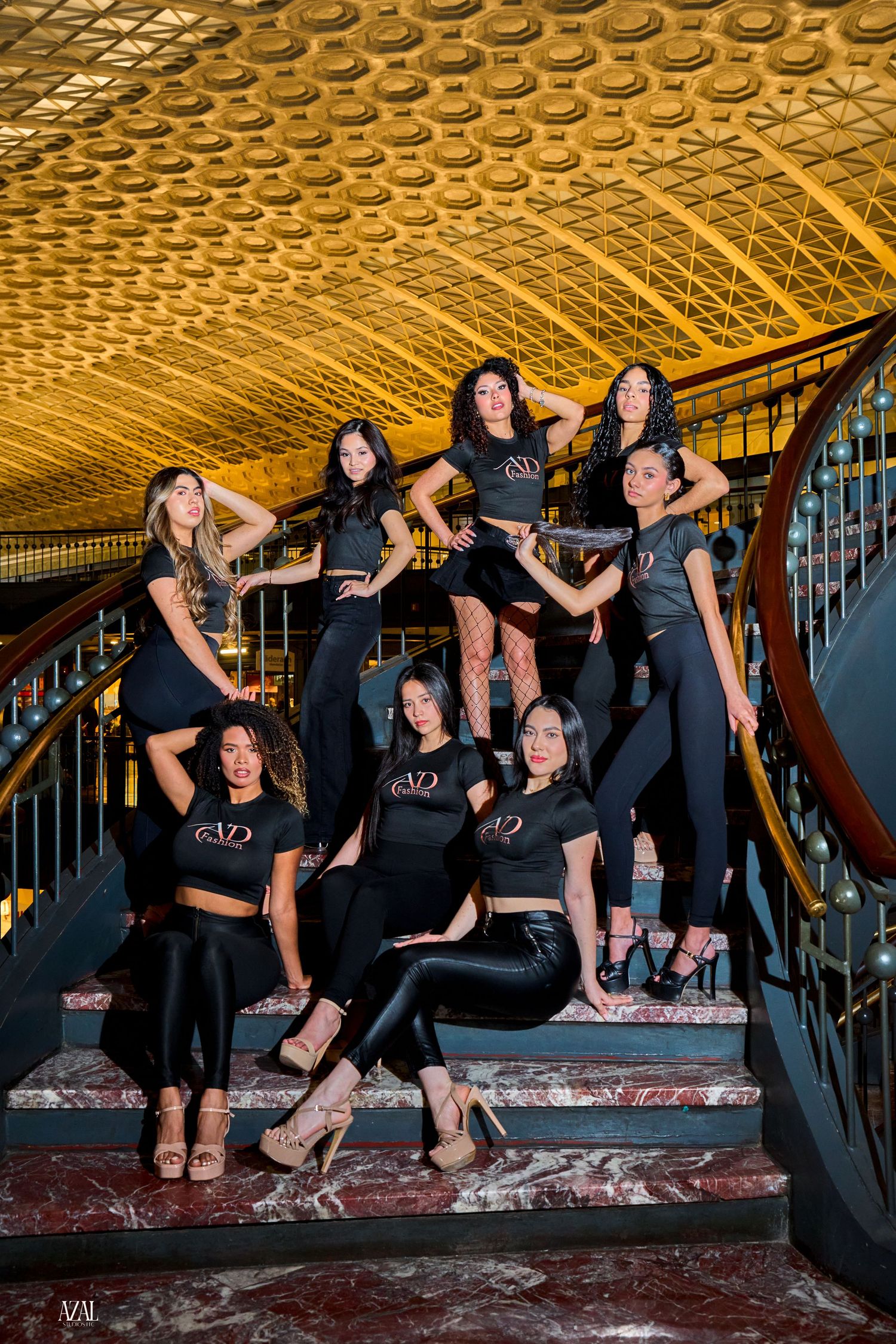 Group of eight women in black VD Fashion outfits pose dynamically on ornate marble staircase.