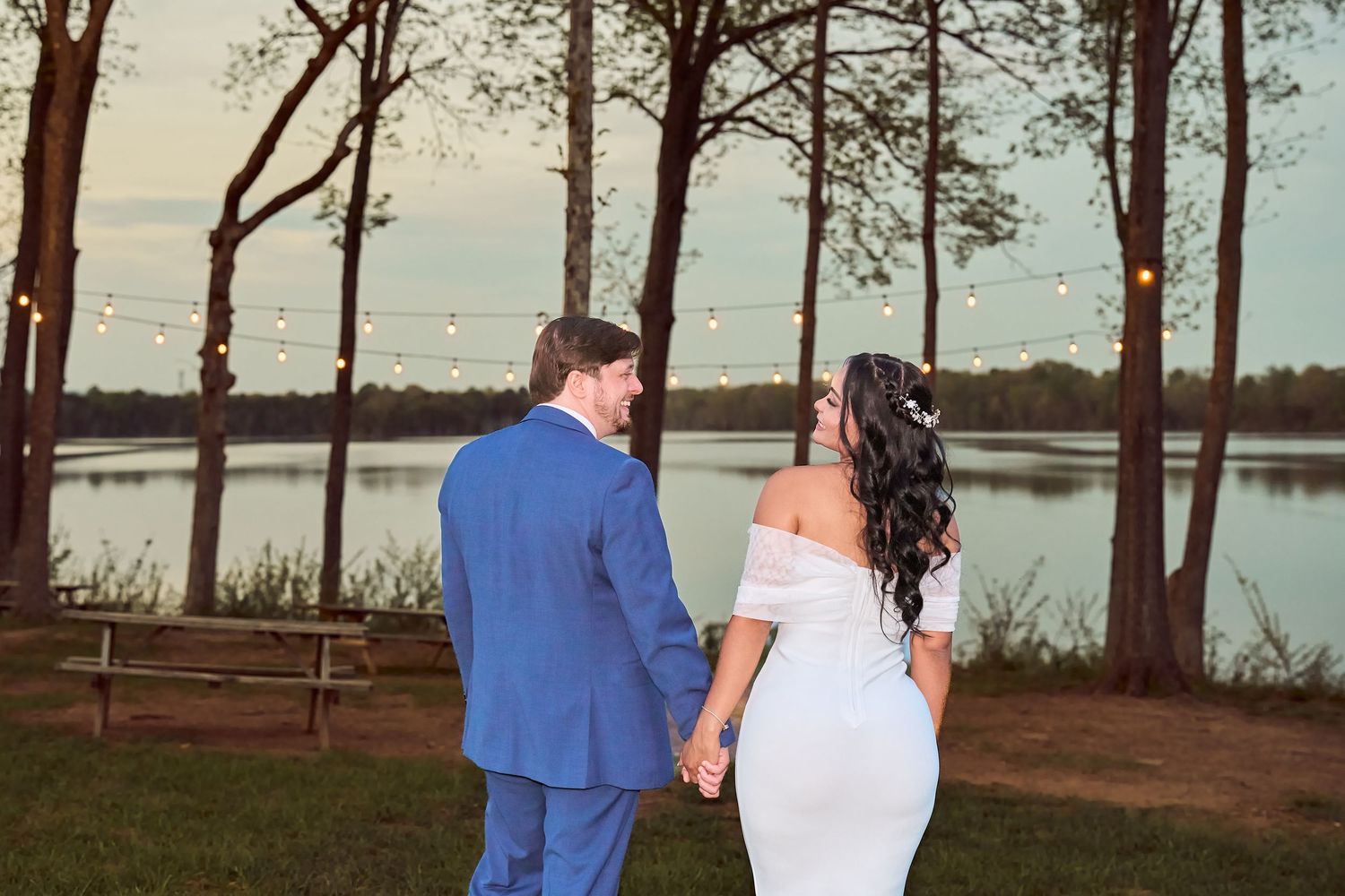 Bride and groom holding hands facing a serene lake at sunset with string lights and trees in background.