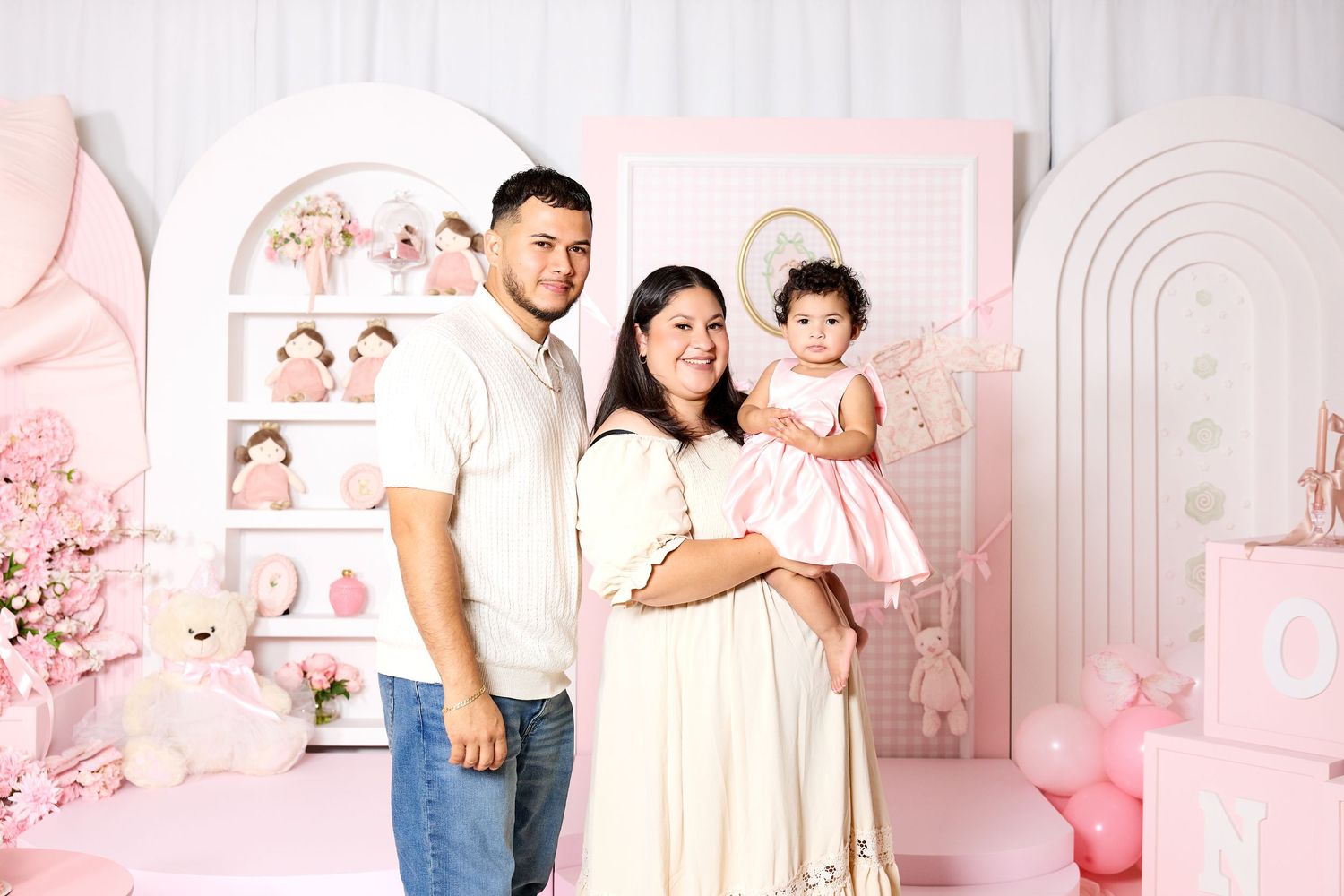 A family of three poses in a pink-themed birthday setup, with parents holding their baby girl in a pink dress.