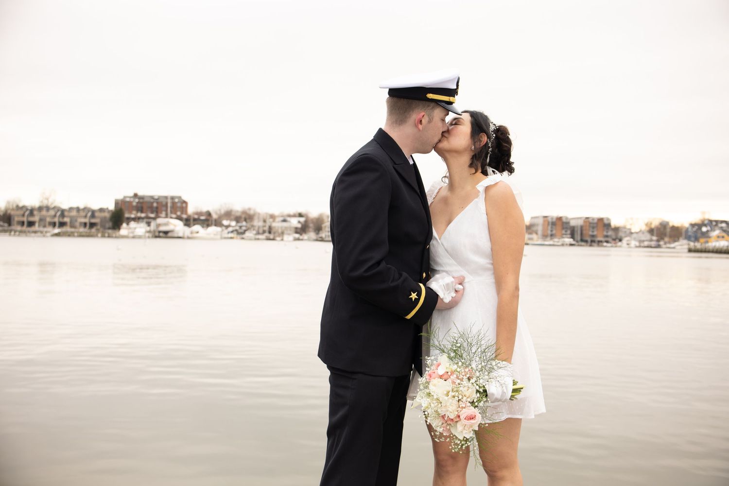 A romantic wedding photo of a couple kissing on a waterfront dock in formal attire.