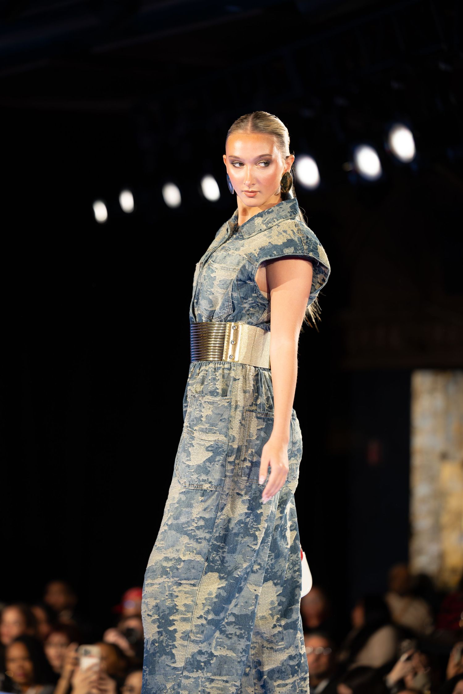 Model wearing military-inspired camouflage dress with metallic belt on fashion runway.