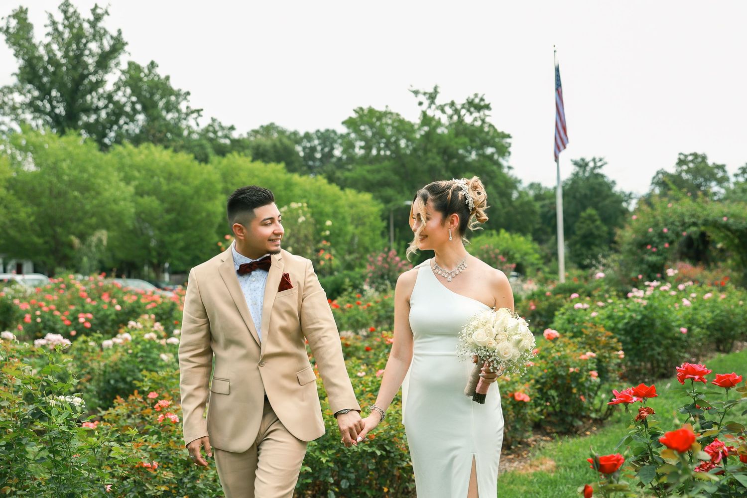 Two people holding hands in a blooming rose garden, wearing formal wedding attire.