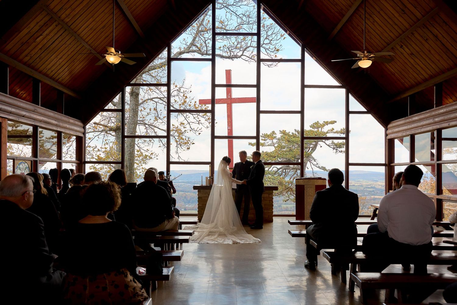 A wedding ceremony in a rustic chapel with large windows and wooden beams featuring a cross on the back wall.