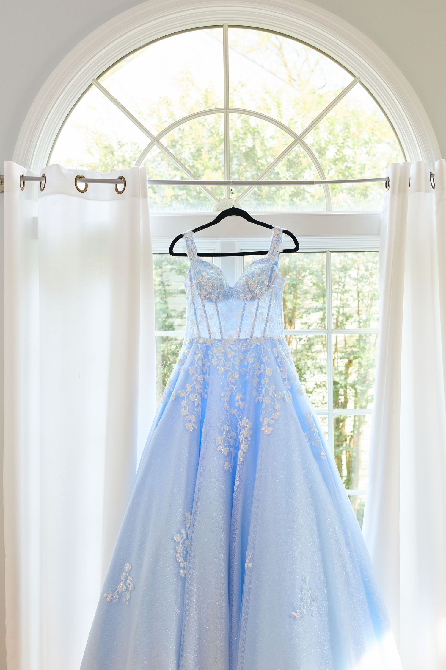 Light blue tulle prom dress with delicate embellishments hanging in a sunlit window display.