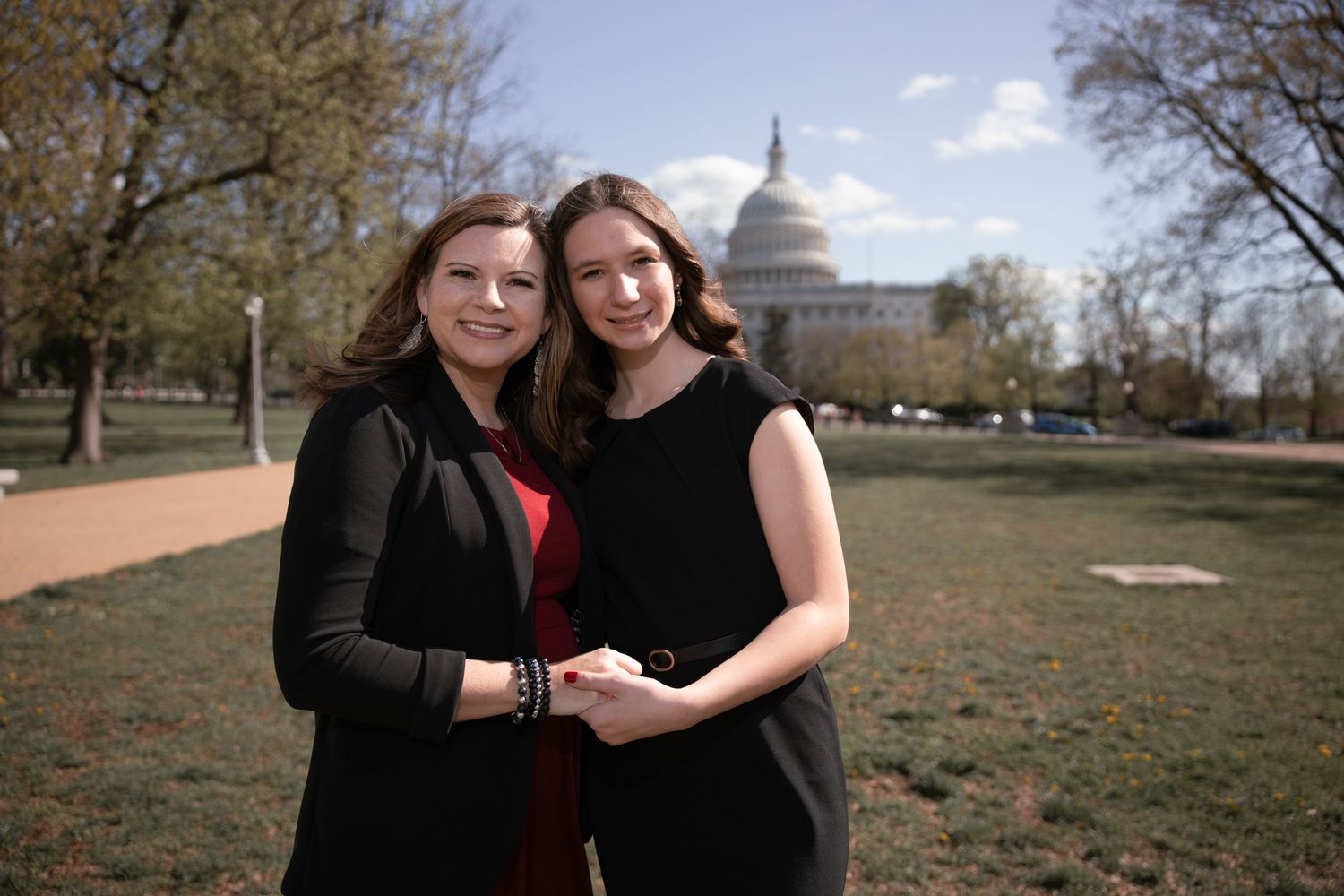 Two people in dark clothing stand together on grassy lawn with US Capitol dome visible in background.