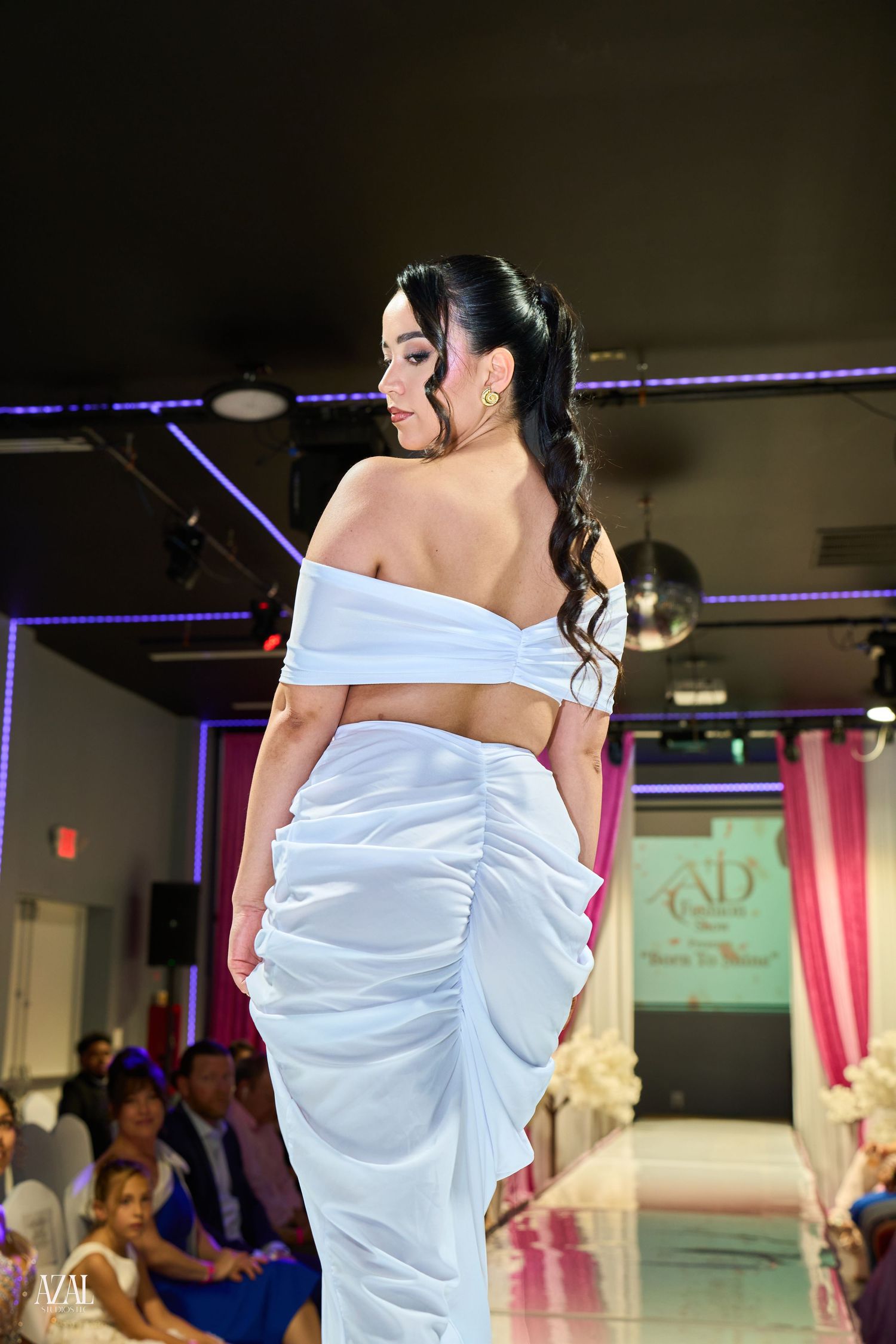 Model on runway wearing elegant white off-shoulder ruched gown at AD Fashion show.