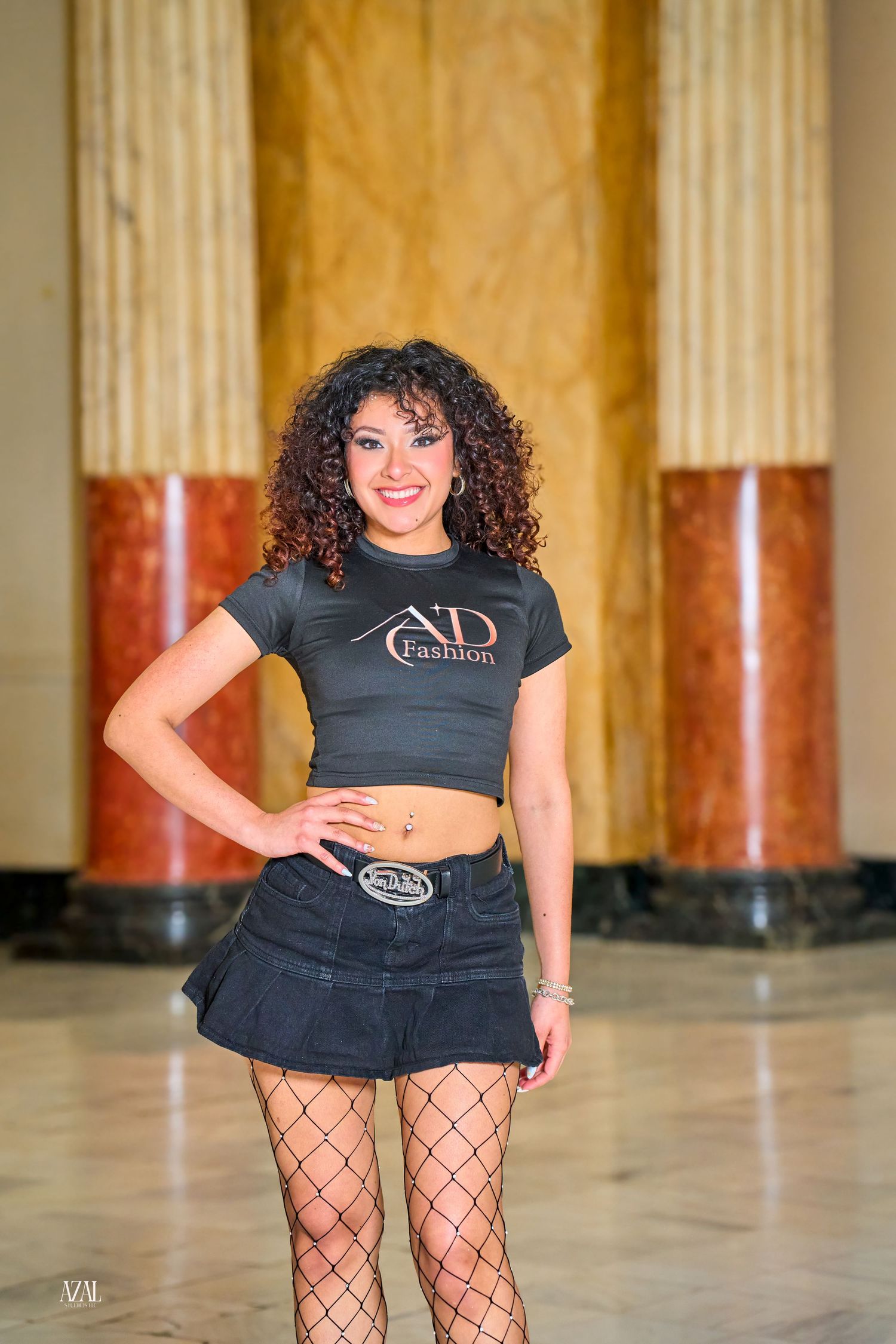 Young woman with curly hair wearing ND Fashion crop top, denim mini skirt, and fishnet tights posing in elegant hall.