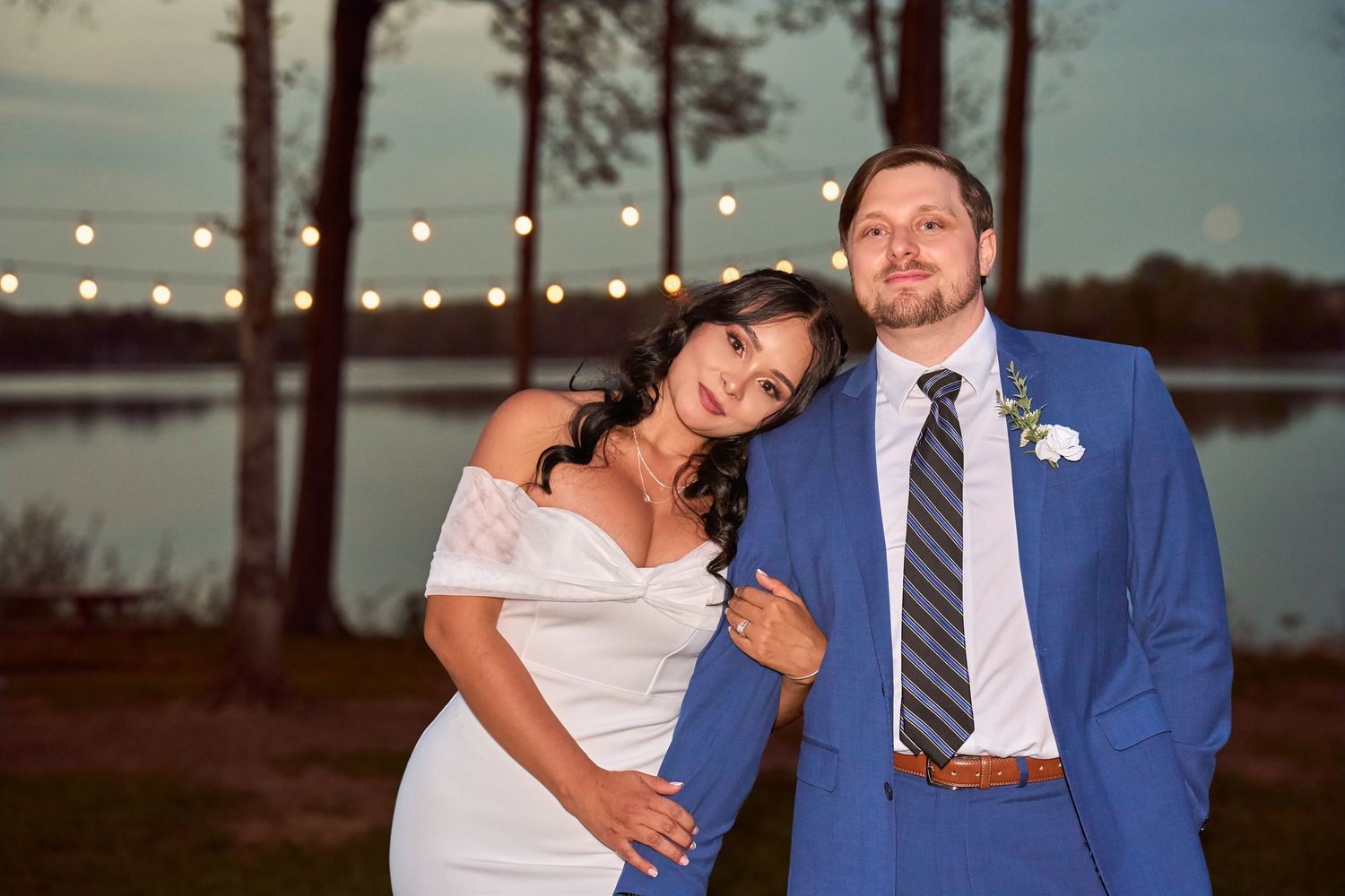 Bride and groom posing outdoors at dusk near a lake with string lights, she in white dress, he in blue suit.