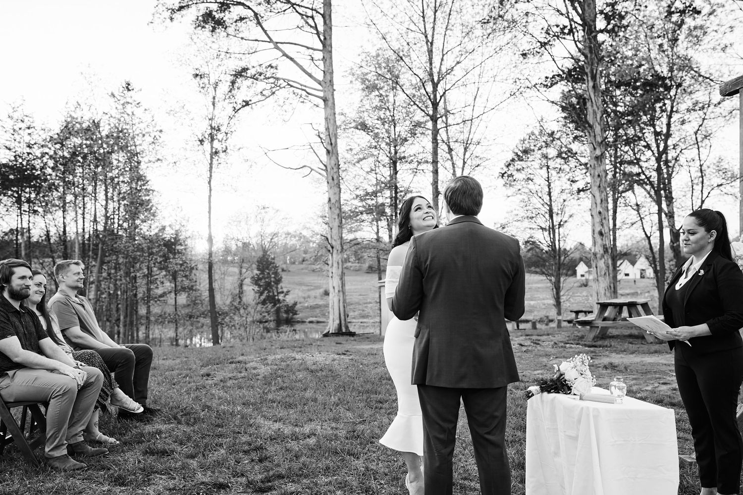 Black and white outdoor wedding ceremony with couple at altar, guests seated on left, officiant on right, trees in background.