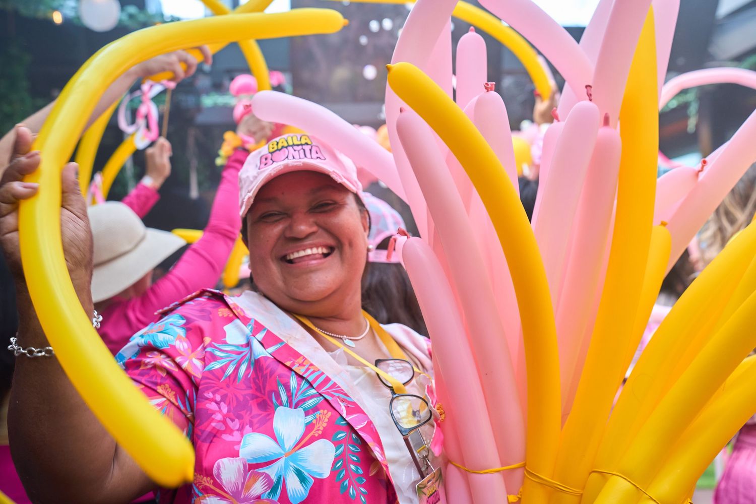 Joyful woman in pink floral outfit holding colorful balloon sticks at a vibrant outdoor festival.