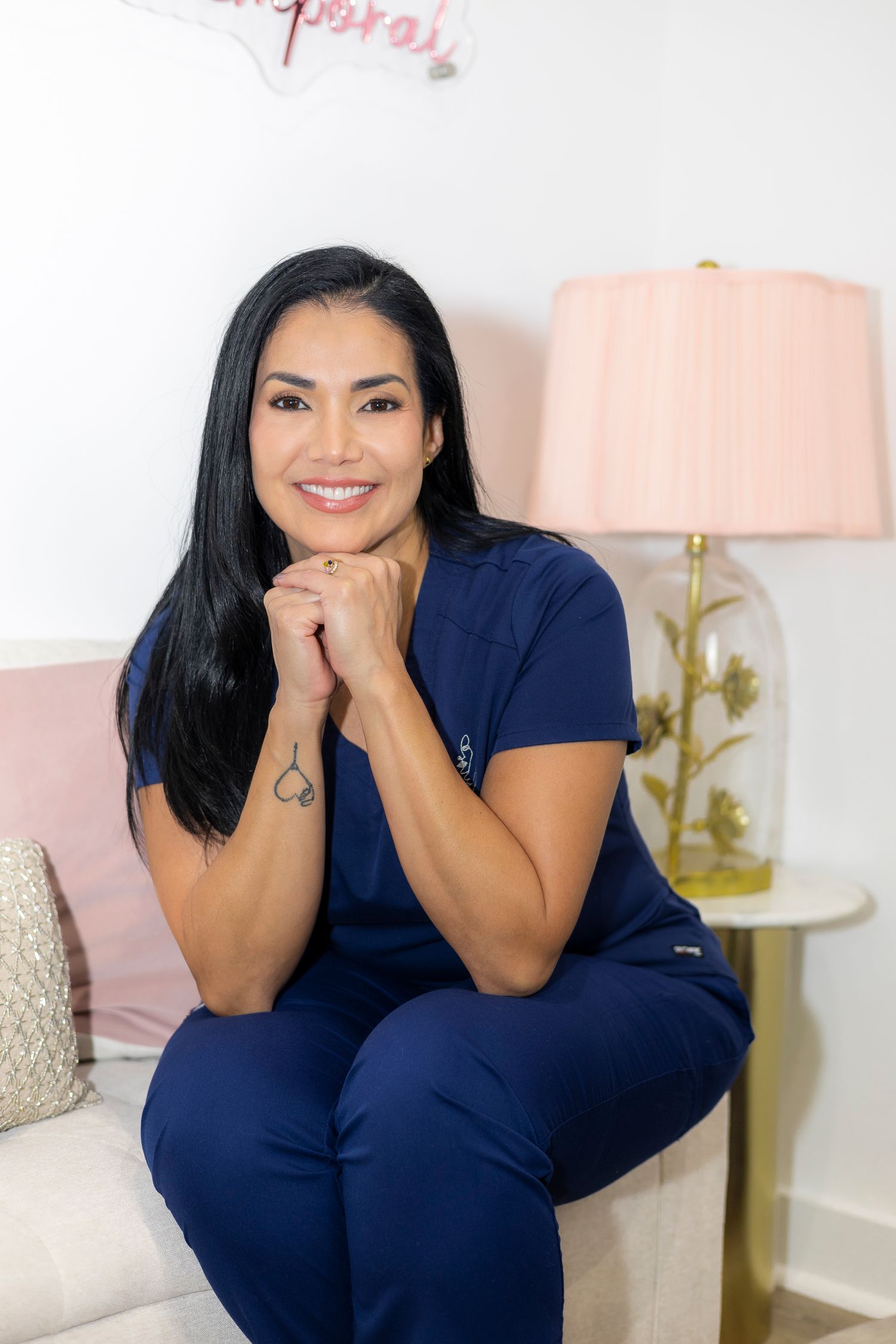 Woman in navy blue medical scrubs sitting on light colored couch with pink lamp in background.