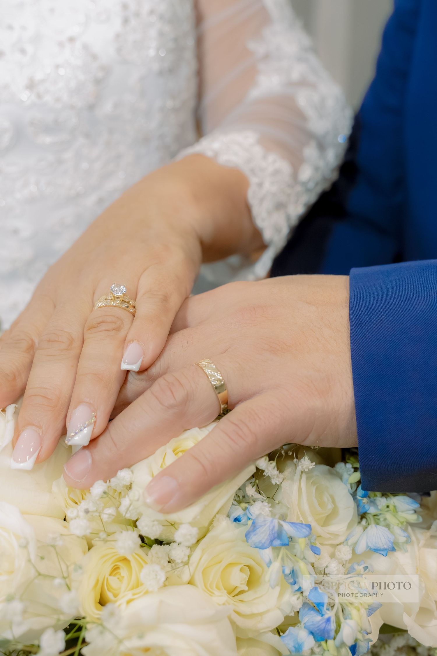 Close-up of wedding rings and manicured hands resting on white and blue flower bouquet.