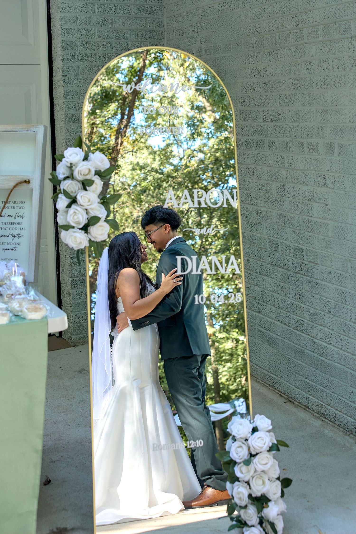 Wedding dress displayed on decorative arch with white floral arrangements against grey brick wall.