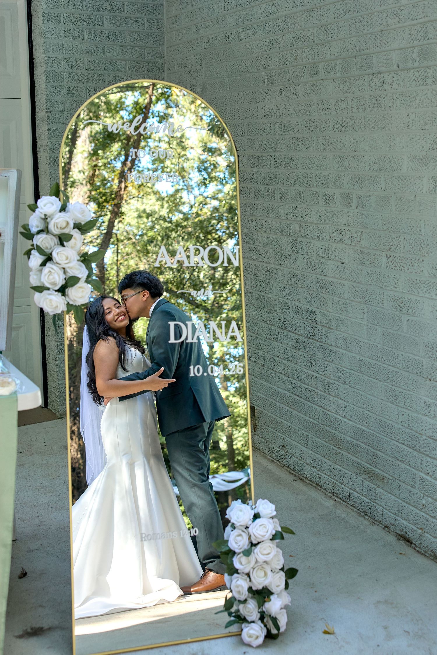 A couple shares a romantic kiss during their wedding ceremony under an archway decorated with white flowers.