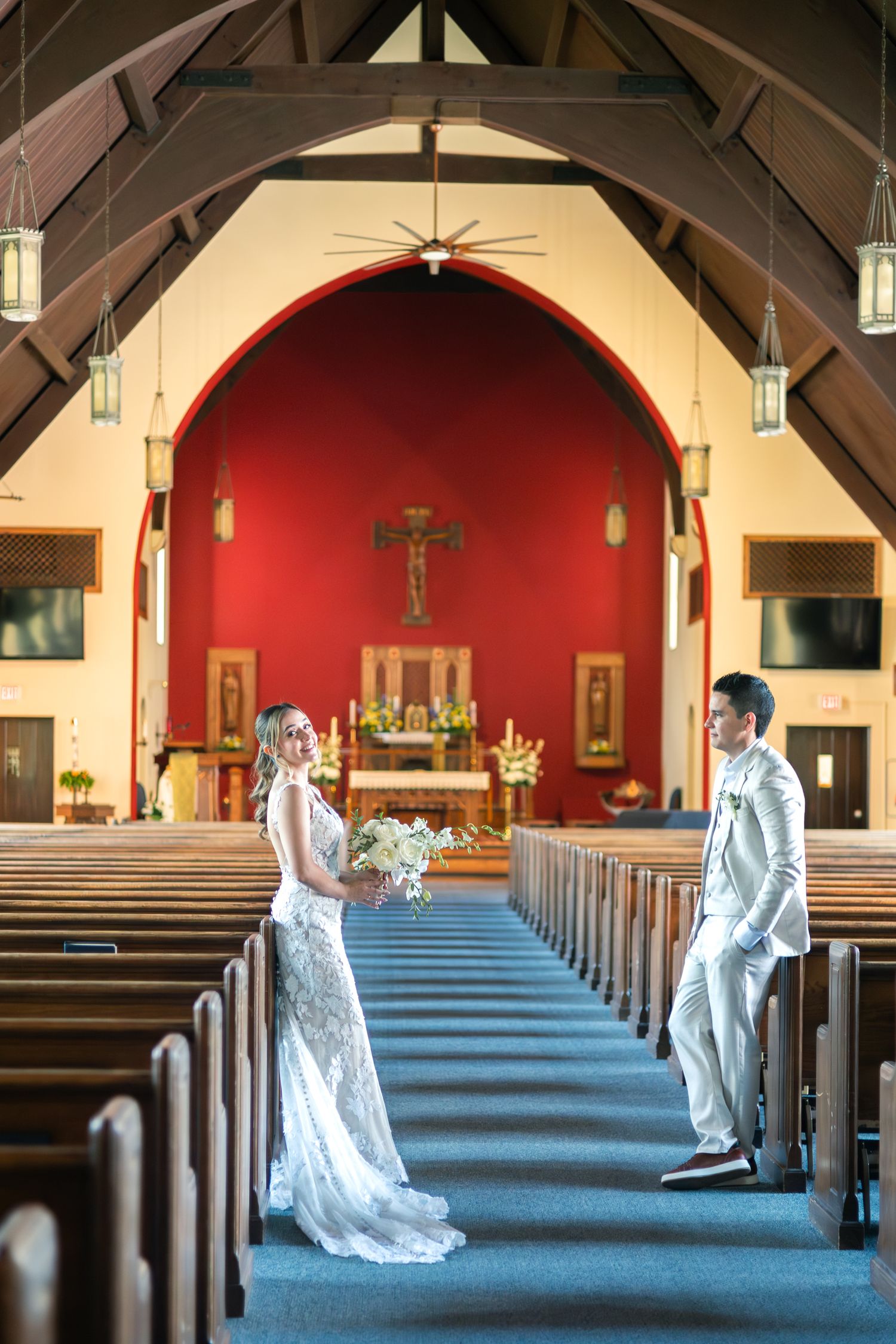 A couple in wedding attire kneels and prays together in the center aisle of a church with red altar walls.