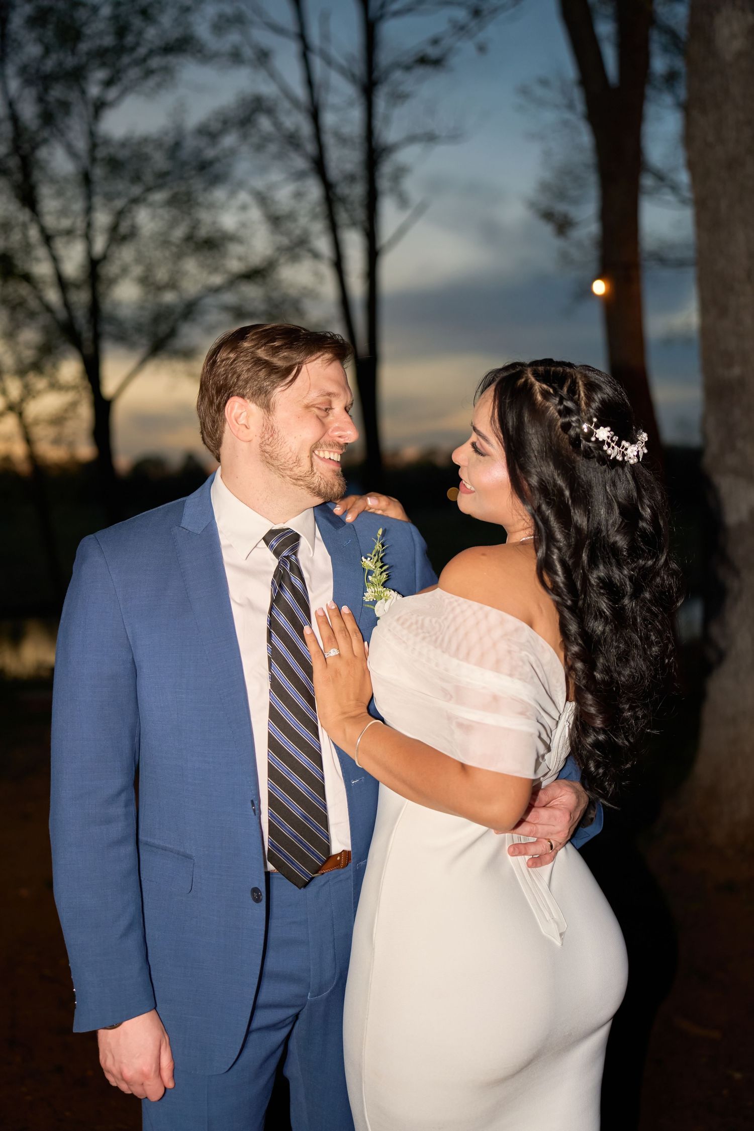 Bride and groom share a romantic moment at sunset, she in a white off-shoulder gown, he in a blue suit with striped tie.