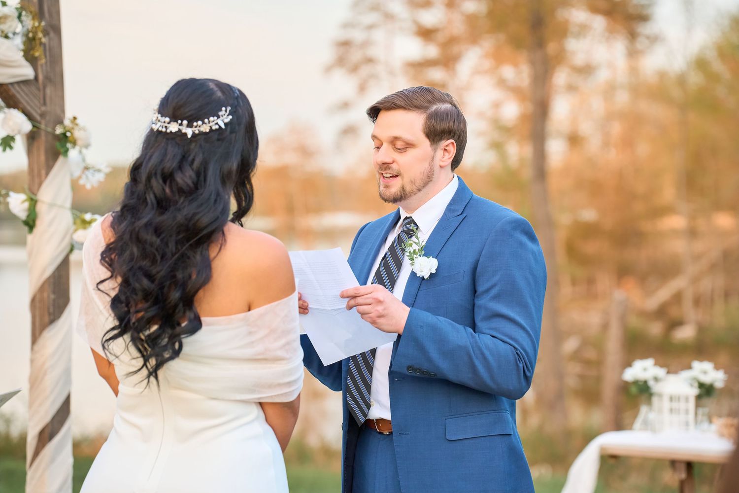 Groom in blue suit reads wedding vows to bride in white off-shoulder dress during outdoor ceremony.