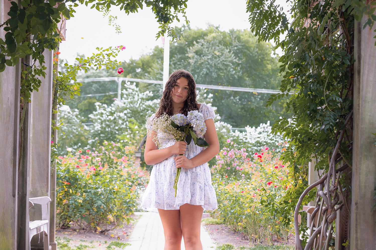 A person in a short white dress poses under a vine-covered archway in a garden with blooming flowers.
