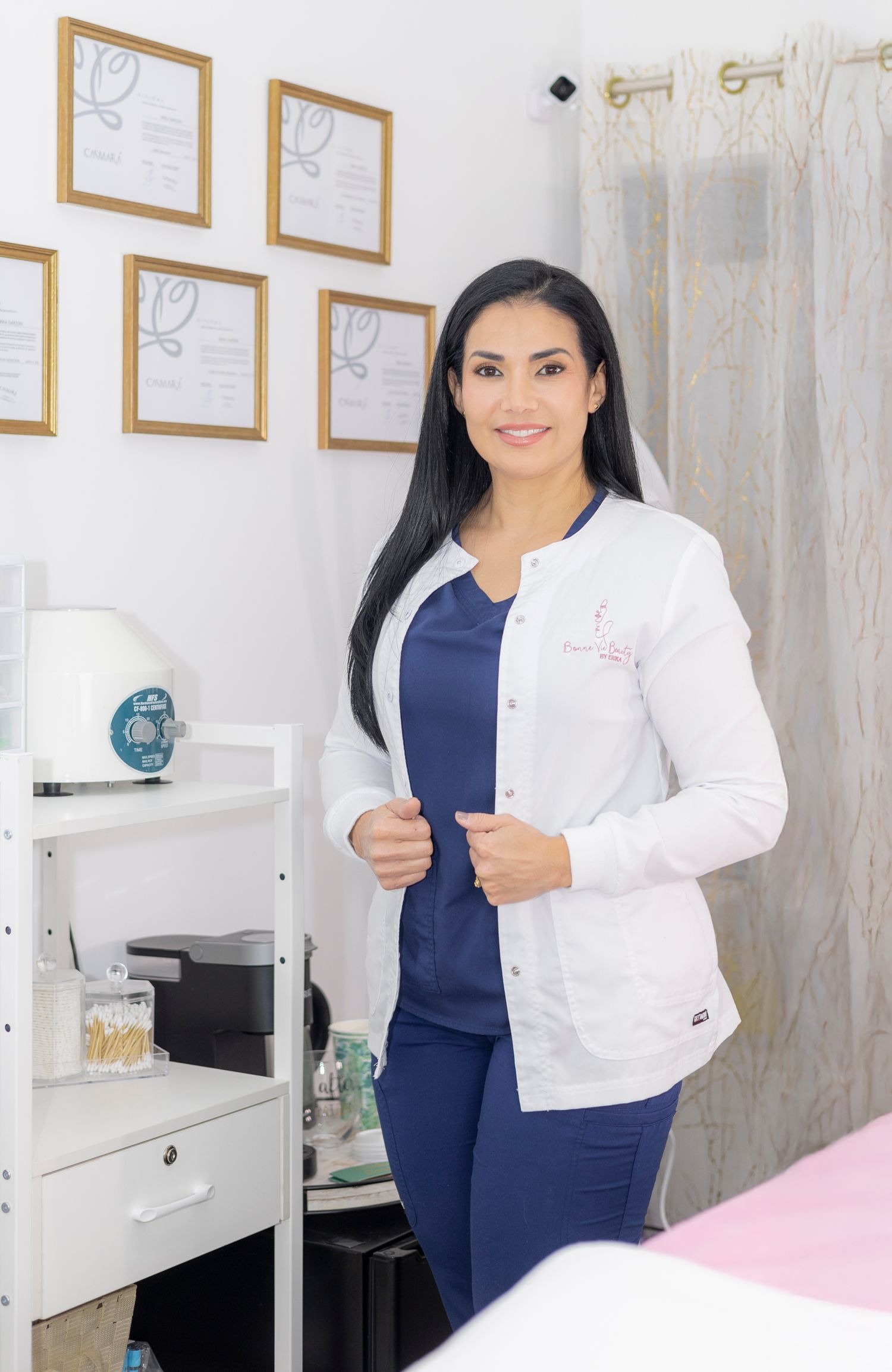 Healthcare professional in white lab coat and navy blue scrubs standing in medical office with framed certificates on wall.