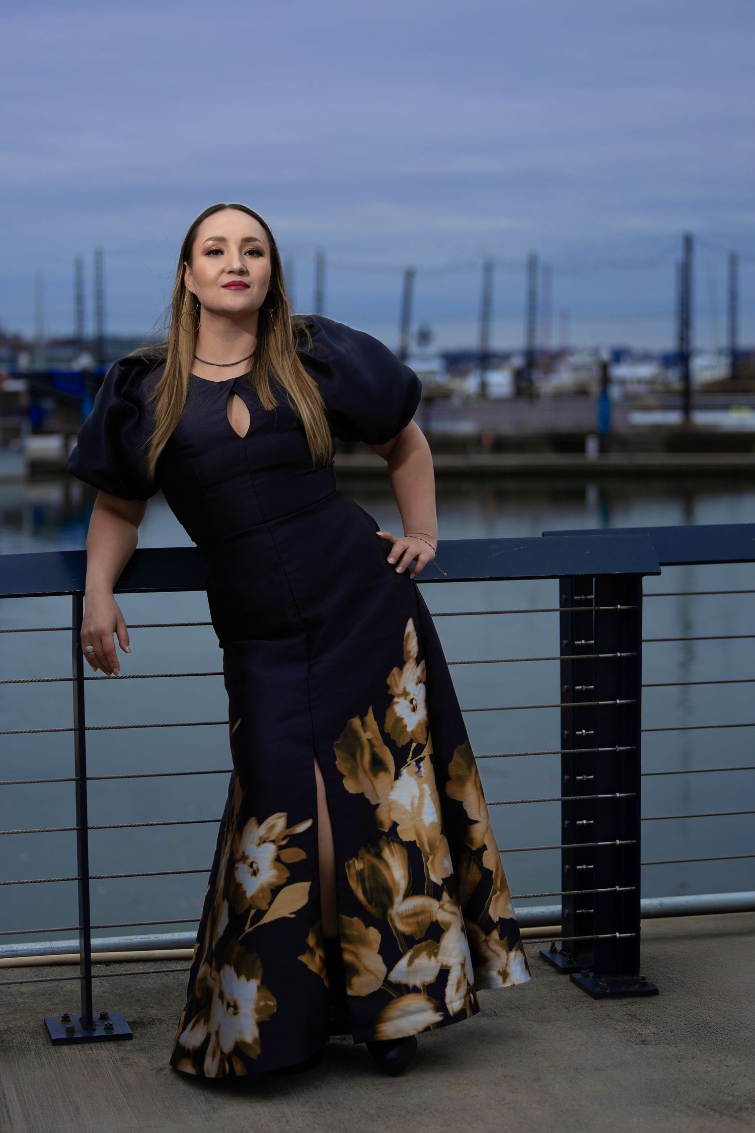 Woman in navy dress with floral print poses on waterfront deck at twilight.