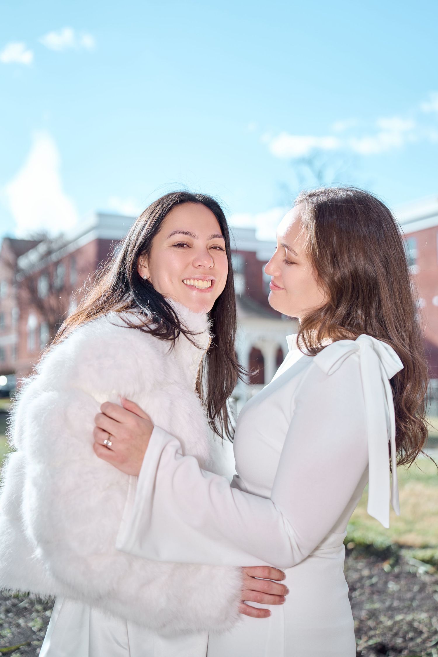 Two people wearing white winter outfits laugh and embrace while standing on a lawn near brick buildings.