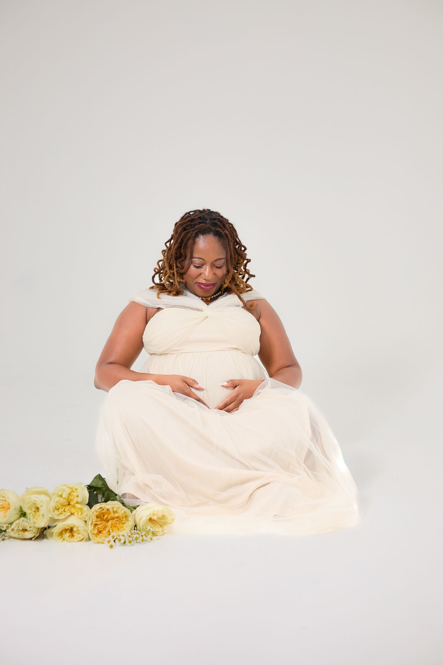 Pregnant Black woman in cream dress cradling baby bump, seated with yellow roses, maternity photo shoot.