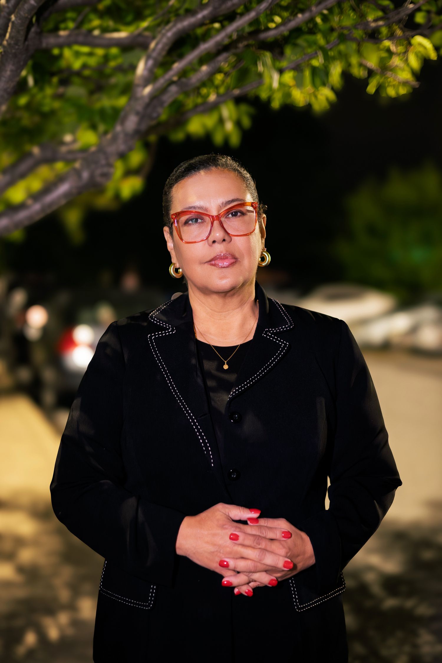A person wearing a black jacket and red glasses poses outdoors with blurred foliage in the background.