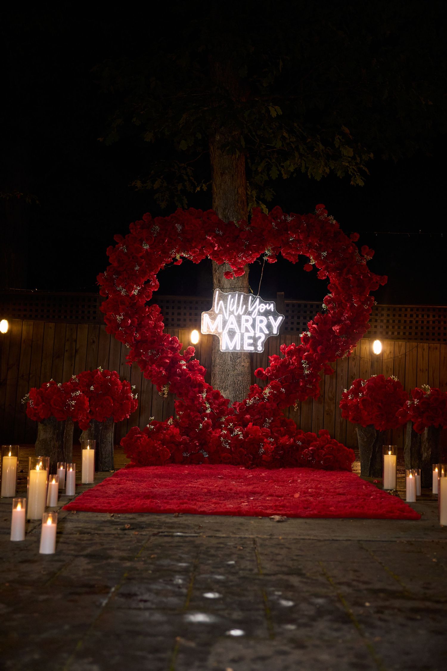 A romantic proposal setup with a heart-shaped red rose arch, neon 'Will You Marry Me?' sign, and lit candles along the path.
