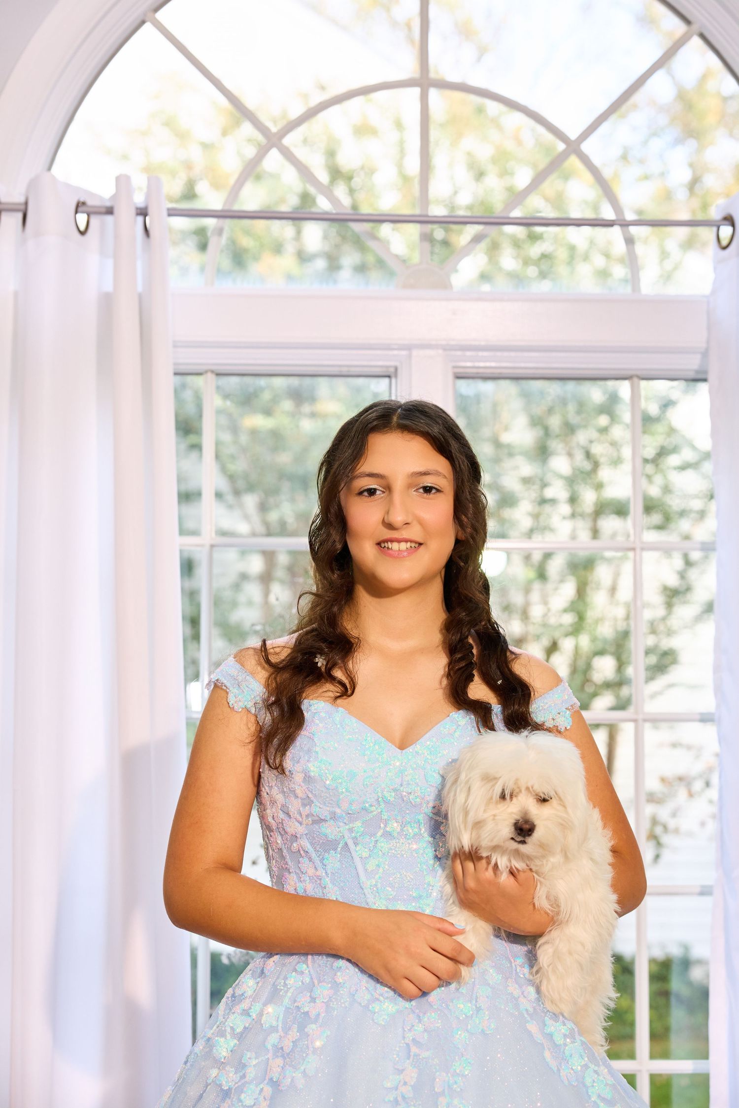 A figure in a light blue dress holds a small white dog while standing in front of bright windows.