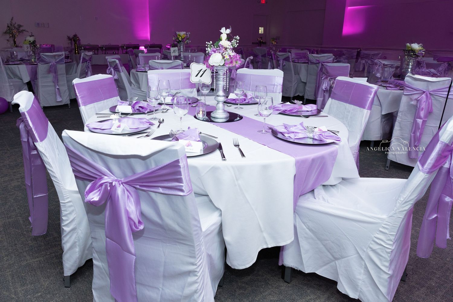Elegant wedding reception table setup with white chairs and purple satin sashes in a dimly lit venue.