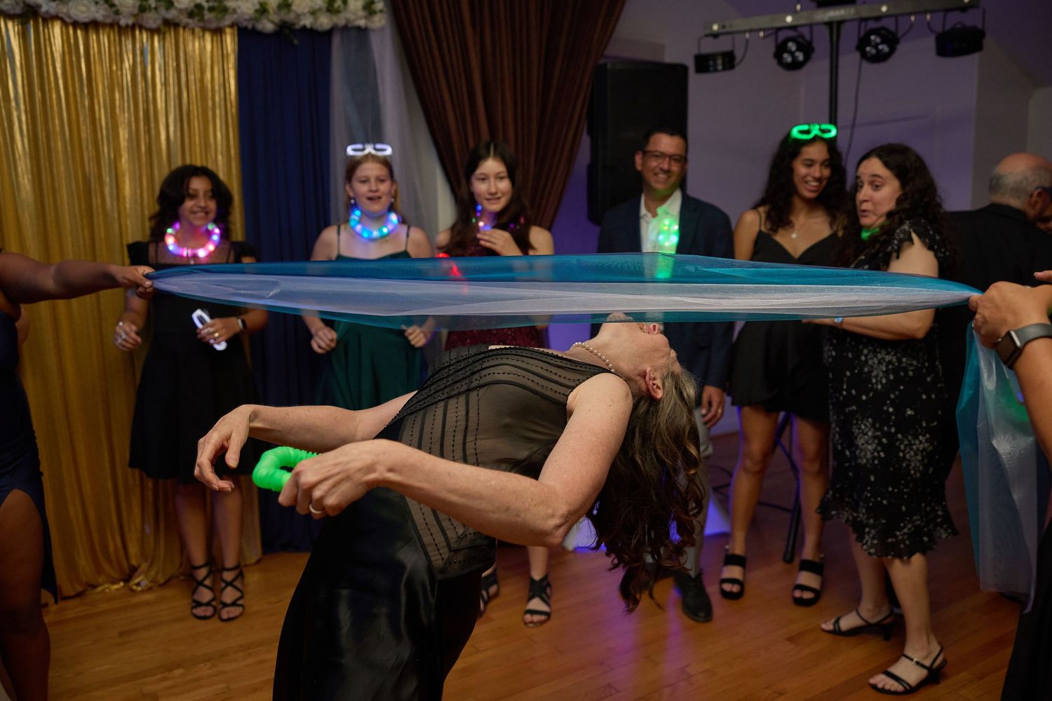 Party guests attempt the limbo dance under a blue bar while others watch and cheer at a festive celebration.