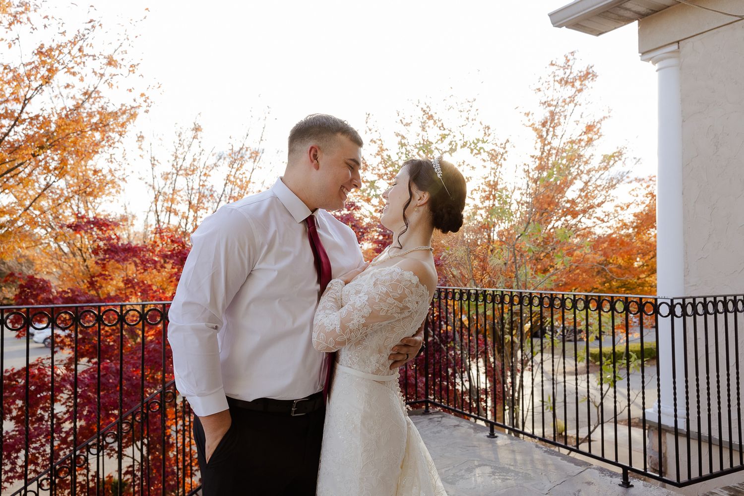 A couple shares a romantic autumn moment on a white porch with vibrant red foliage in the background.
