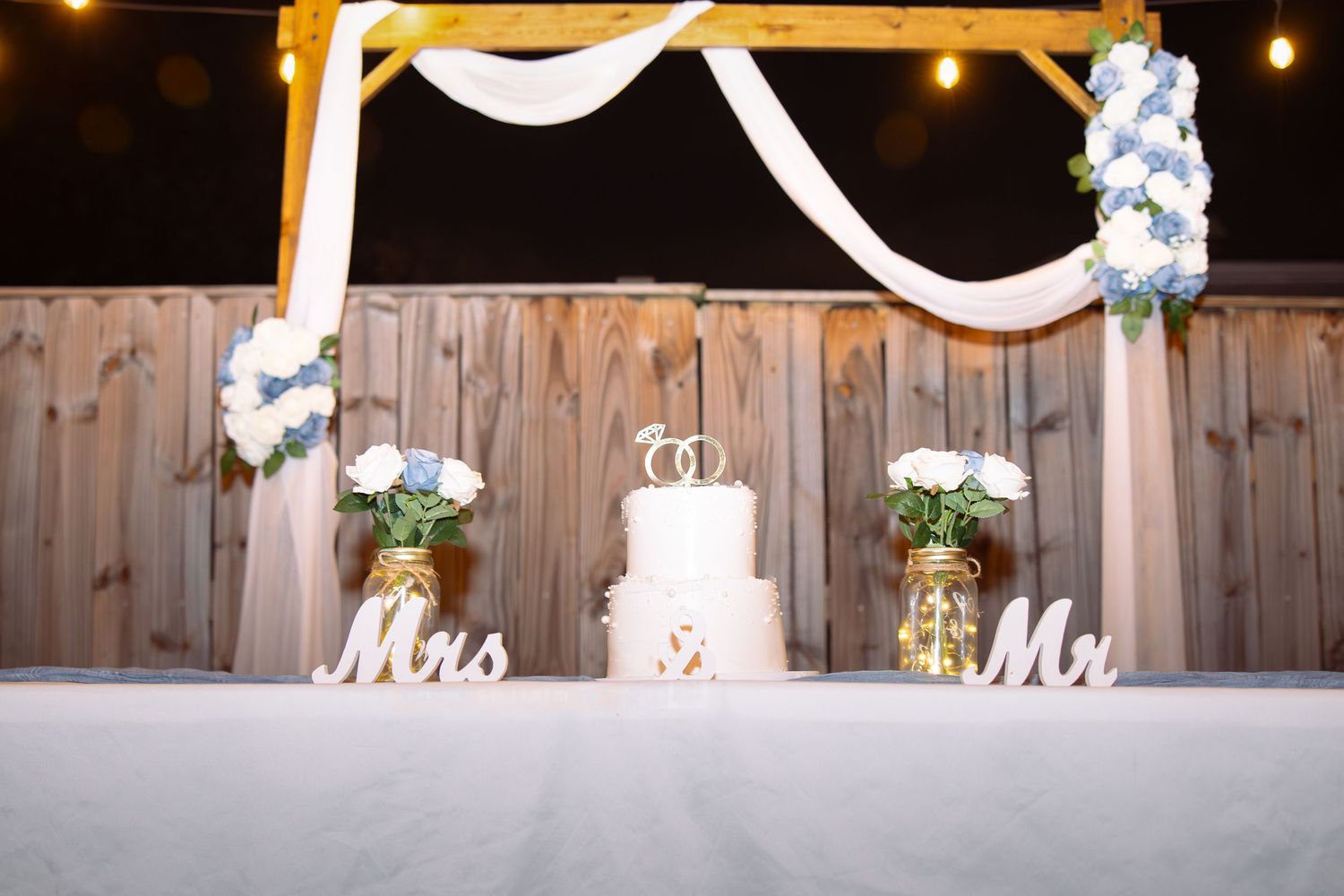 A rustic wooden wedding ceremony setup with white fabric draping and floral decorations under warm string lights.
