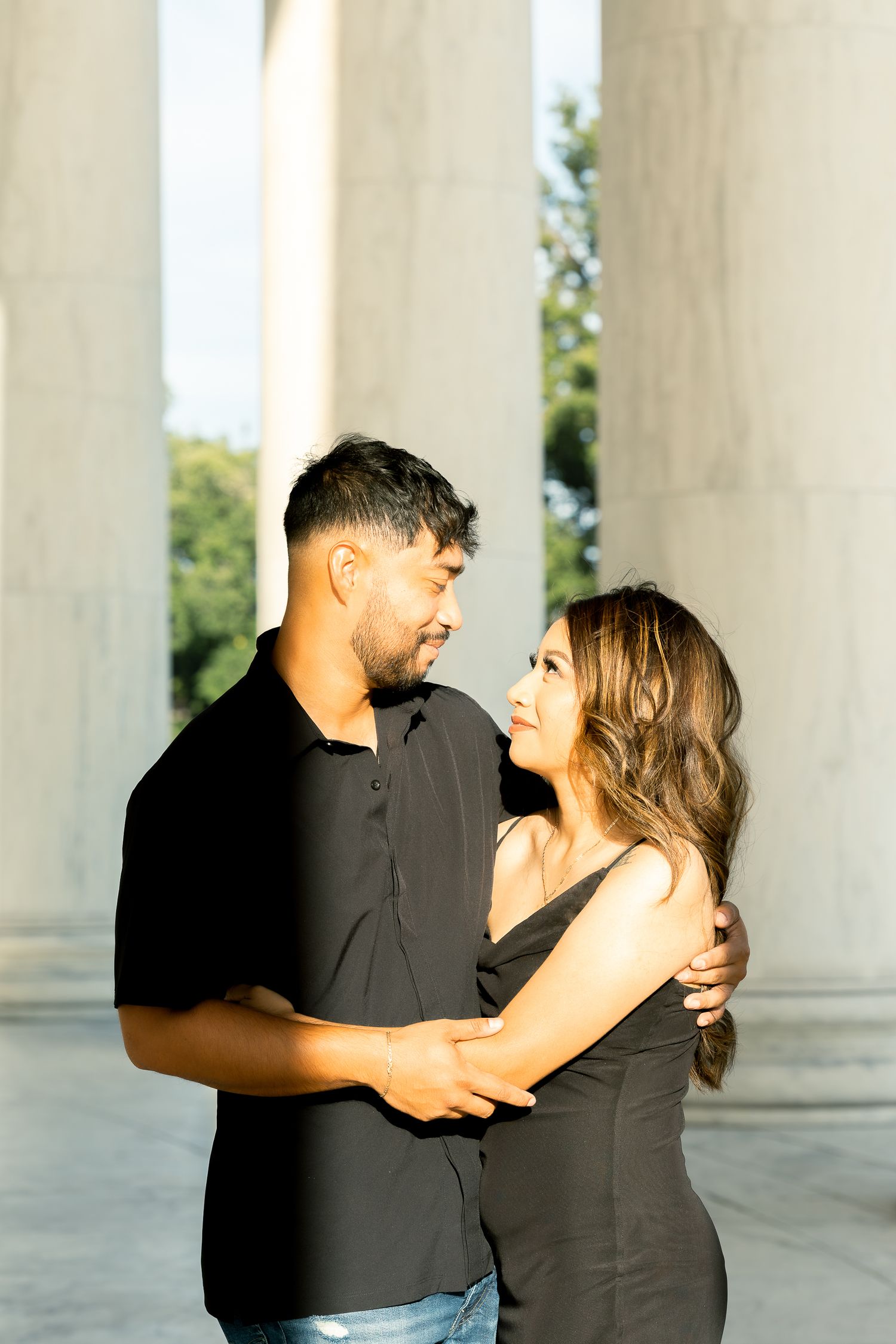 A couple in black outfits share a romantic dance moment between white marble columns in natural sunlight.
