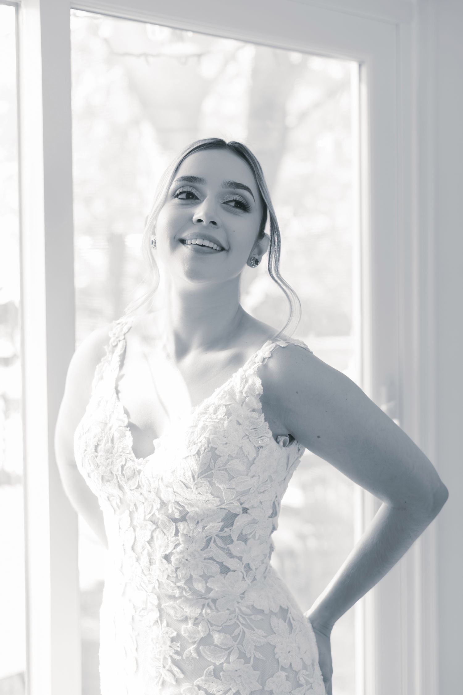 A person in a white lace wedding dress poses elegantly by a window in a bright, ethereal setting.