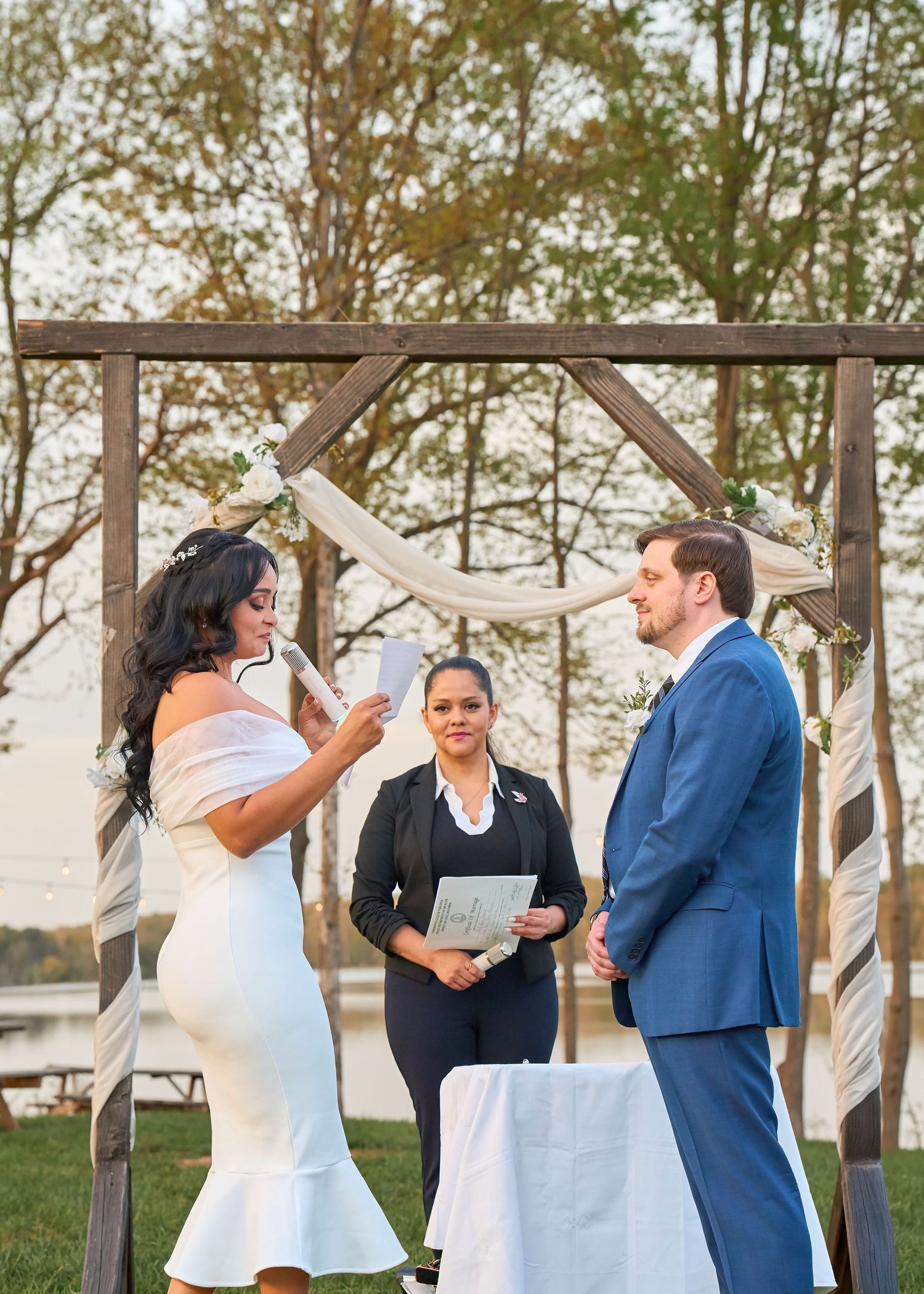 Outdoor wedding ceremony with bride reading vows, groom in blue suit, and officiant under rustic wooden arch.