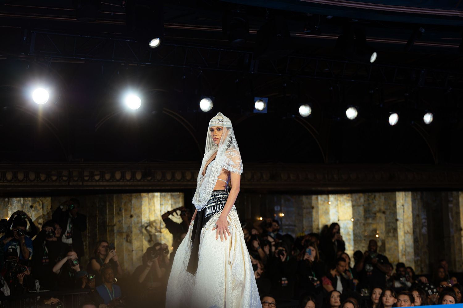 Fashion model in flowing white bridal gown poses dramatically under spotlight on runway.