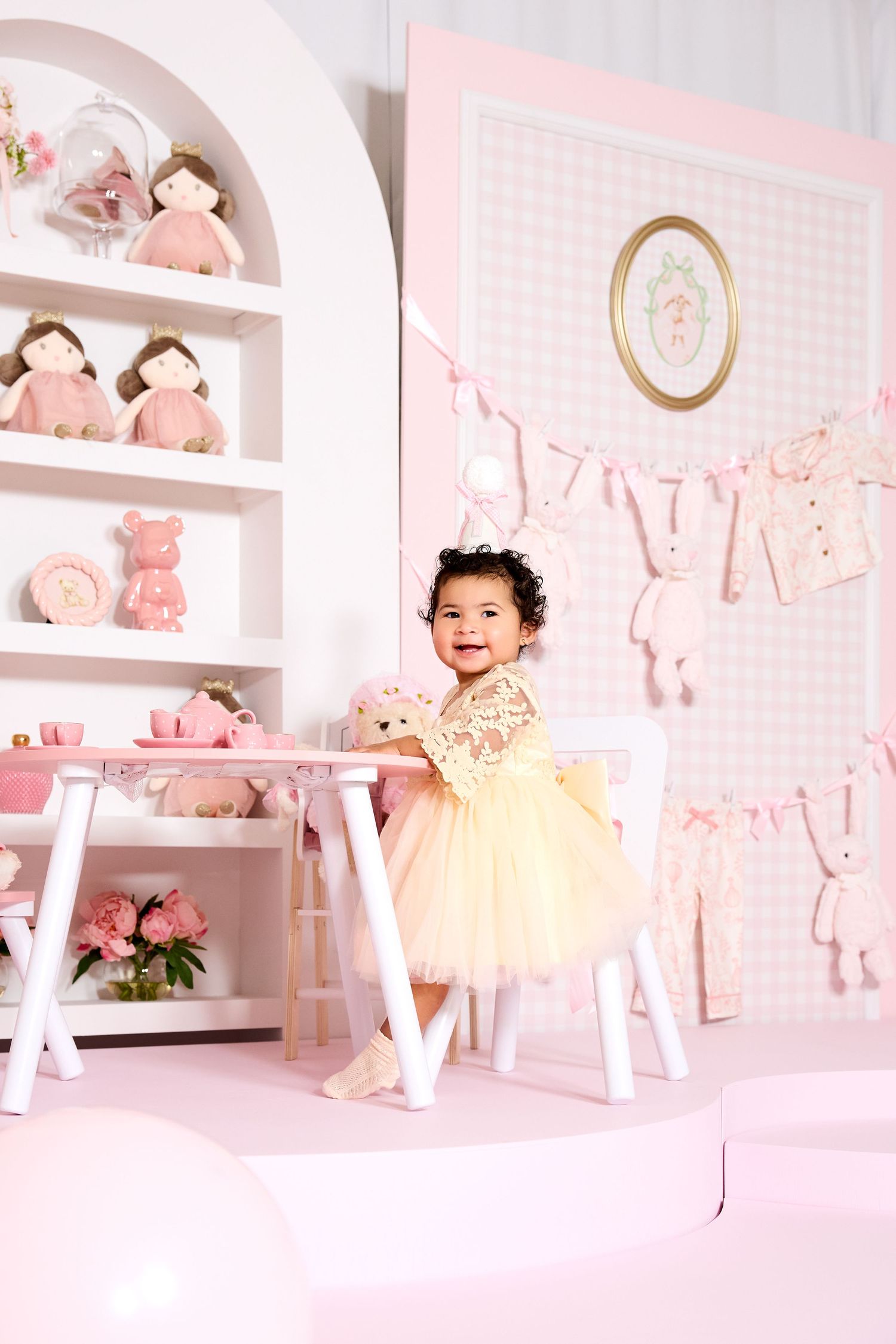 Toddler girl in yellow tutu dress smiling in pink nursery with dolls, bunny decor, and mini table setup.