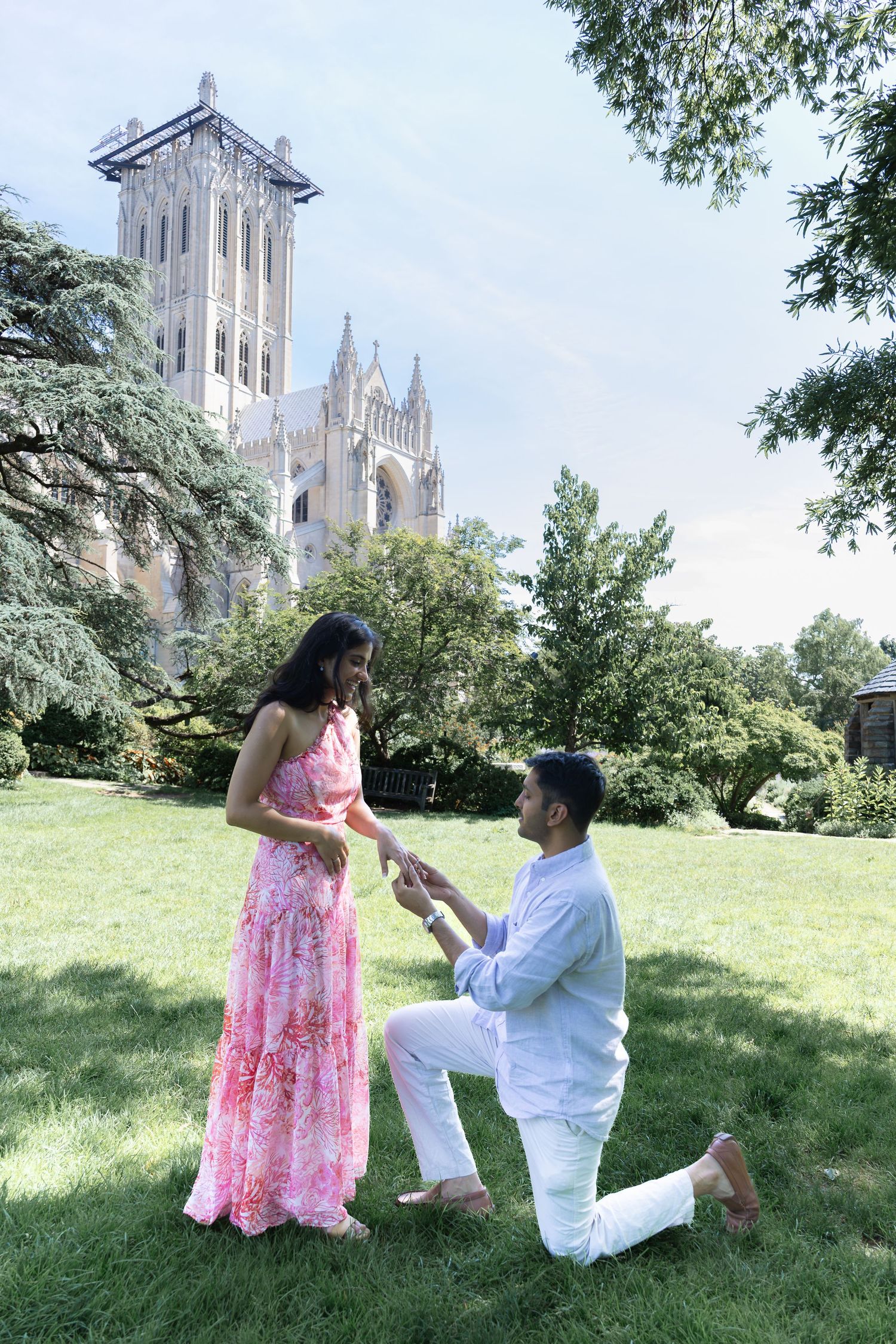 A tender marriage proposal moment unfolds on a grassy hill with a historic cathedral spire visible in the background.