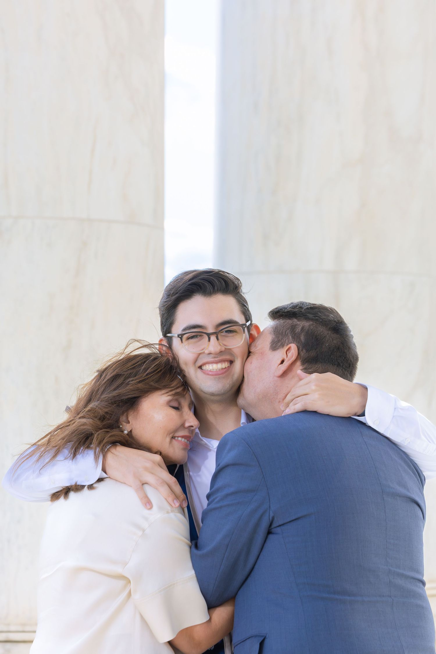 Three people share a warm embrace while laughing together outdoors near a white marble column.