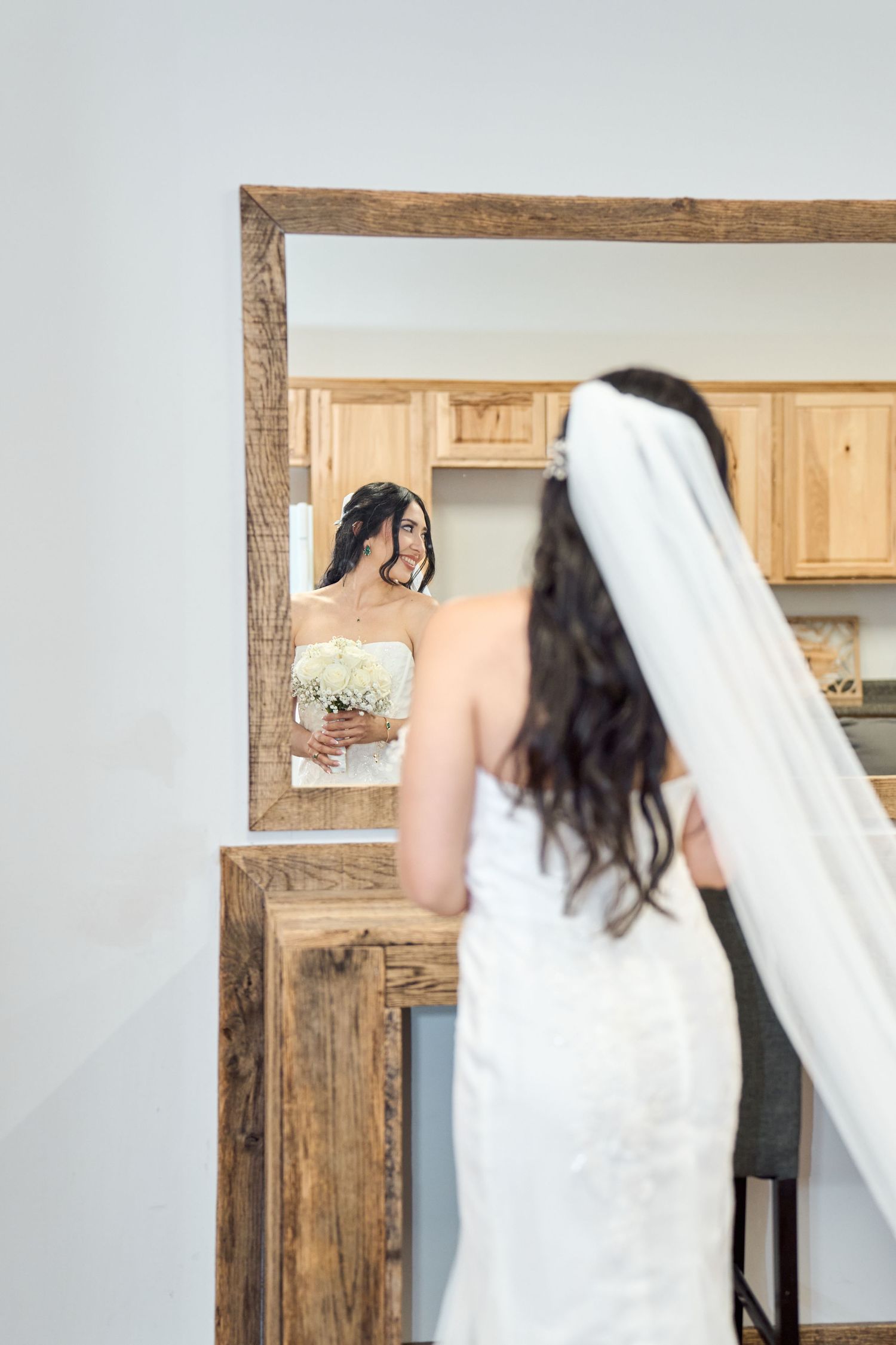 A bride with a long veil looks at her reflection in a rustic framed mirror.