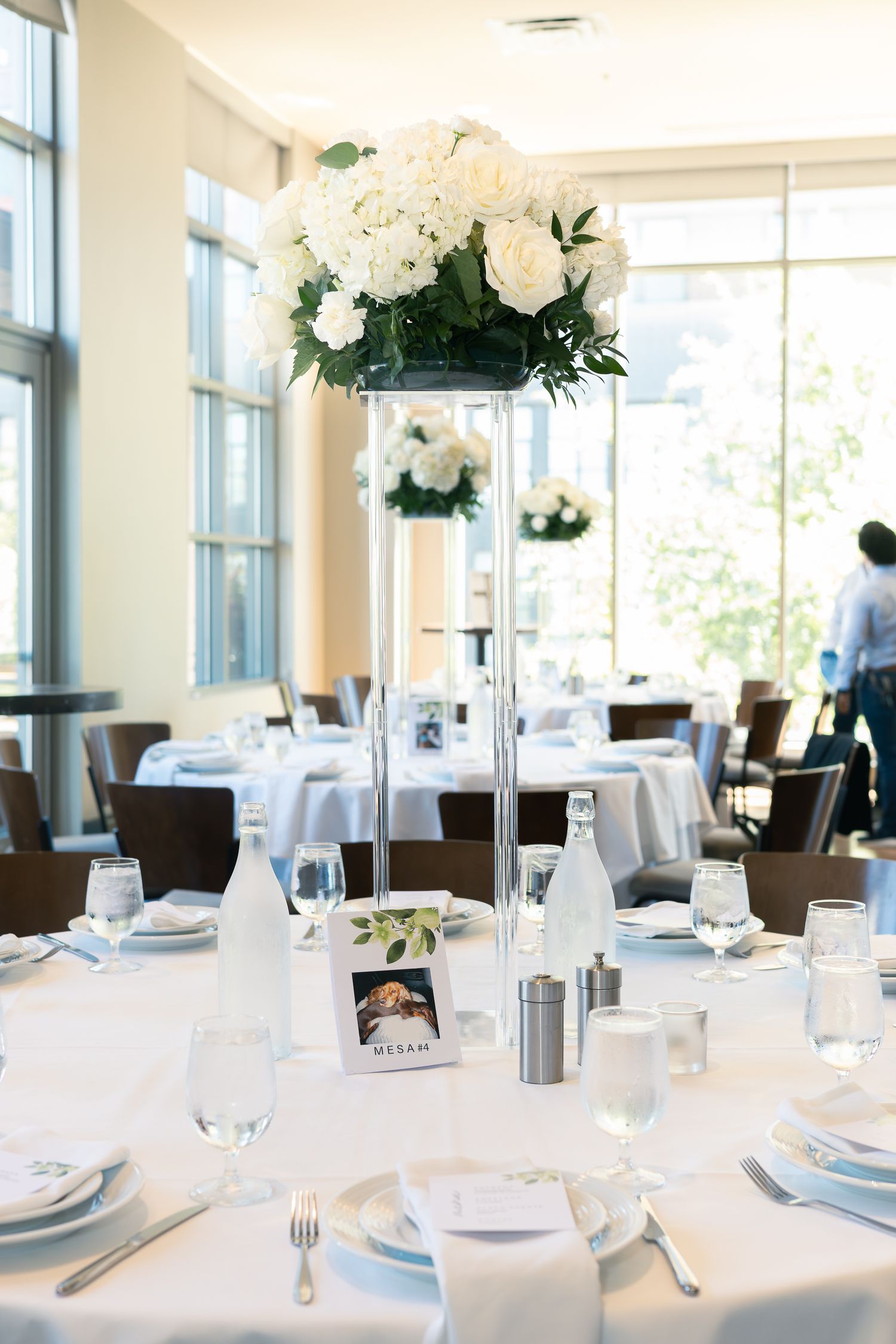 Elegant wedding reception tables set with white linens, tall floral centerpieces and formal place settings in bright sunlit venue.