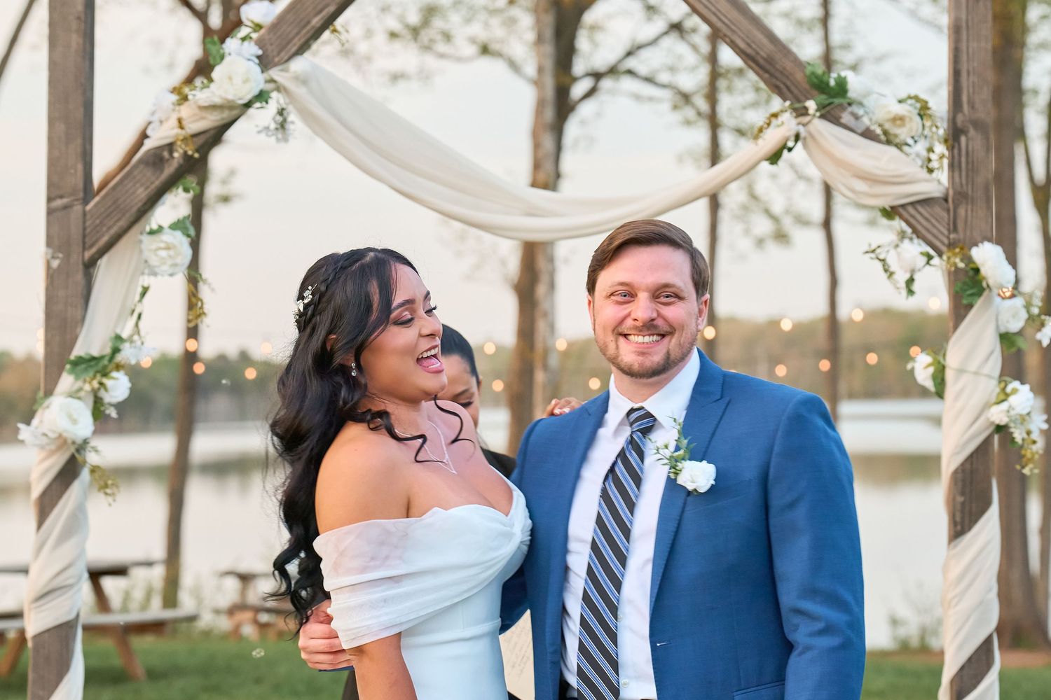 Smiling bride and groom pose under a floral wooden arch by a lake at their outdoor wedding ceremony.