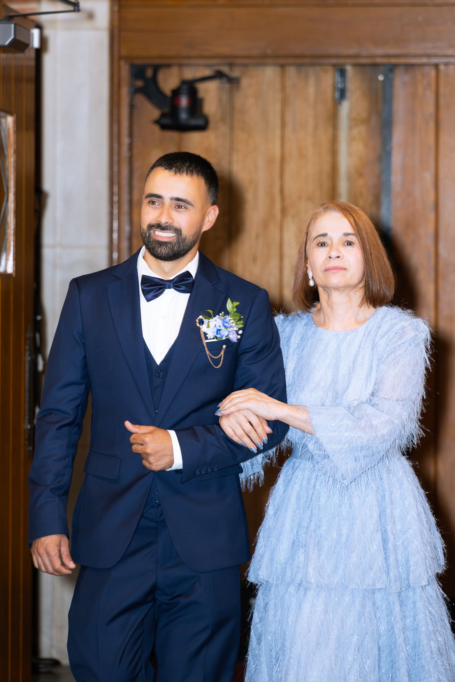 A couple in elegant attire walk arm in arm, with the groom in navy tux and bride in a light blue feathered dress.