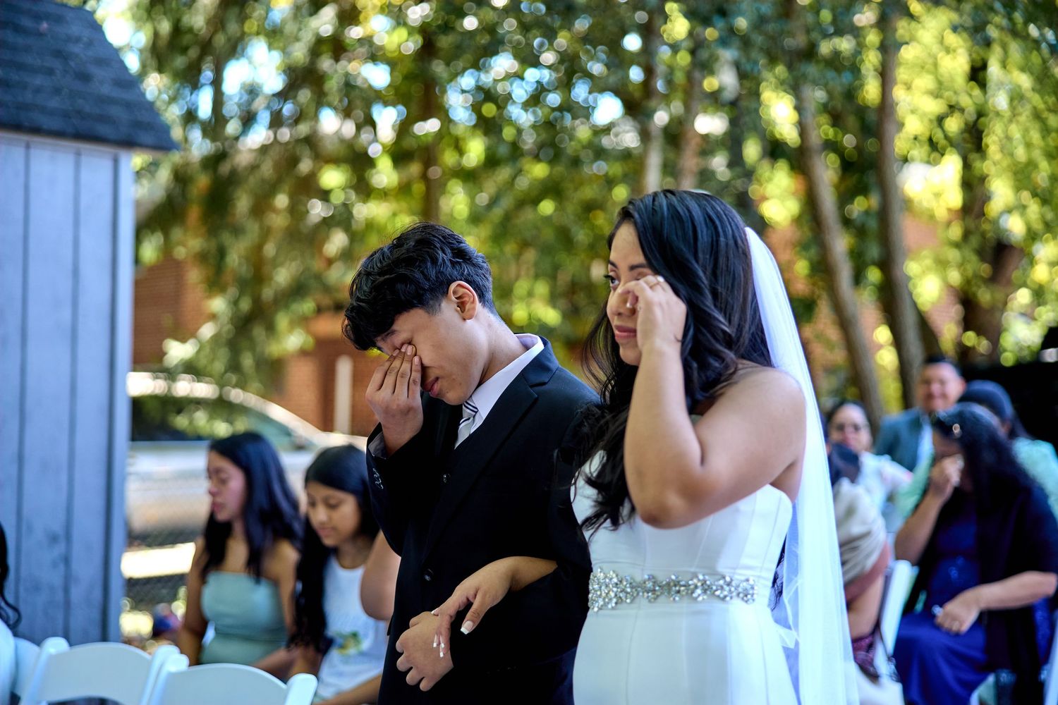 Wedding guests gather outdoors under trees during a daytime ceremony celebration.