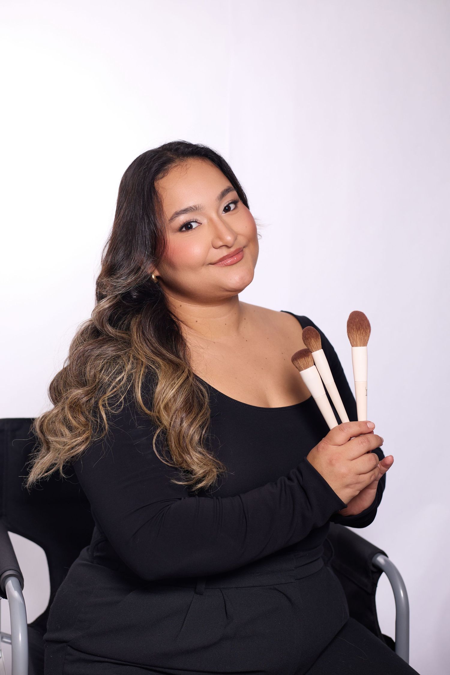 Makeup artist in black sweater poses with brushes against white background in professional photoshoot series.
