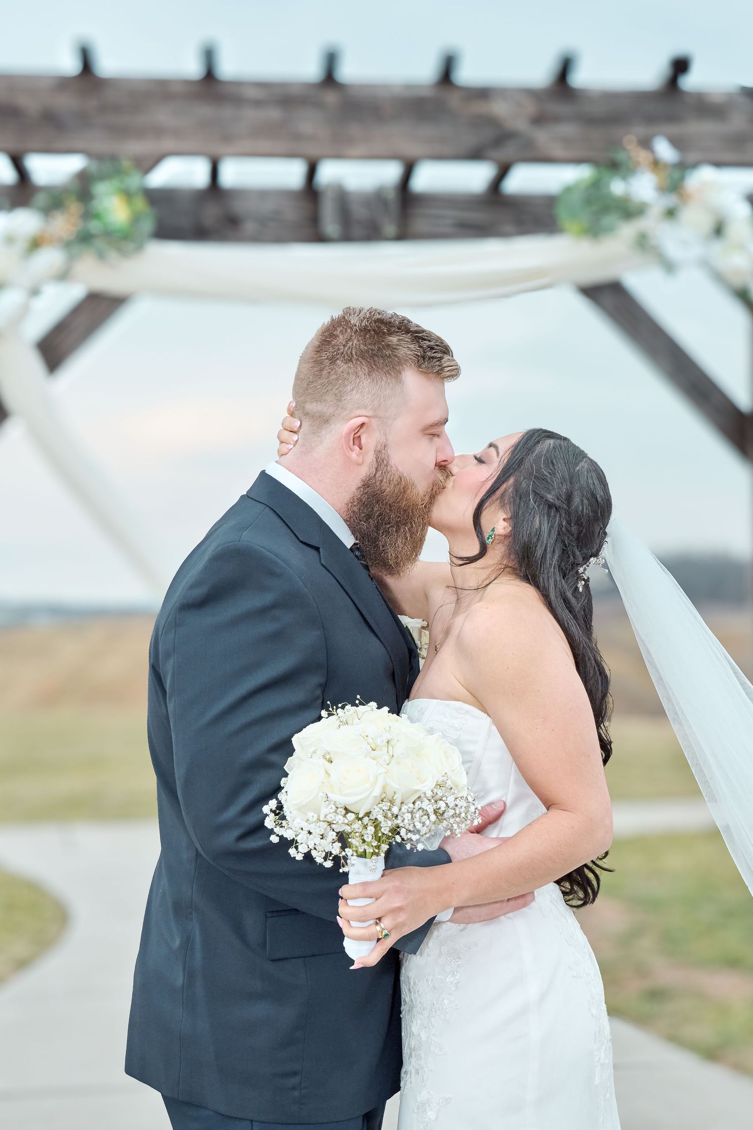 A romantic kiss between newlyweds under an outdoor pergola with white floral decorations.