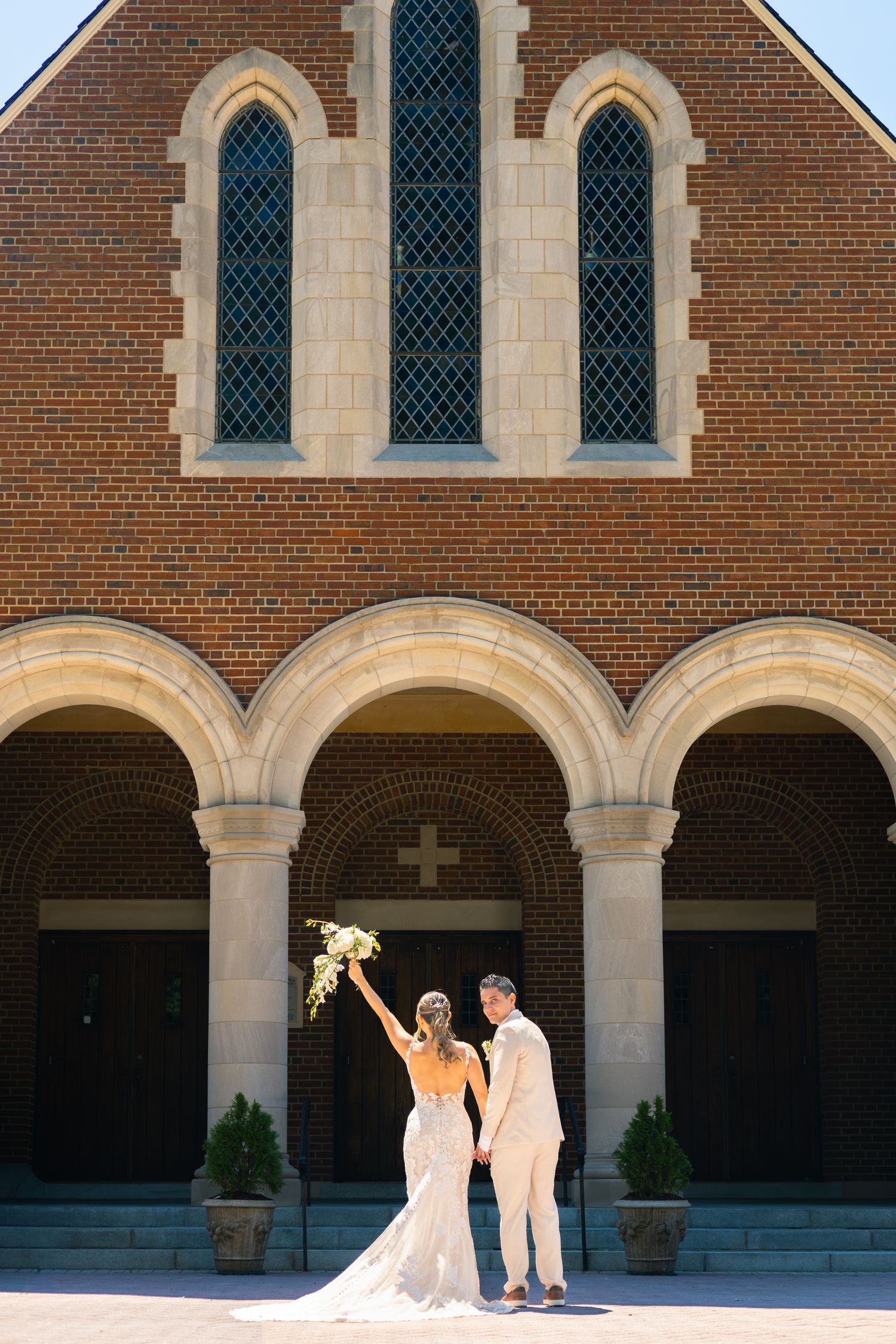 Wedding couple stands under arched brick colonnade with sunlight streaming through the architectural details.