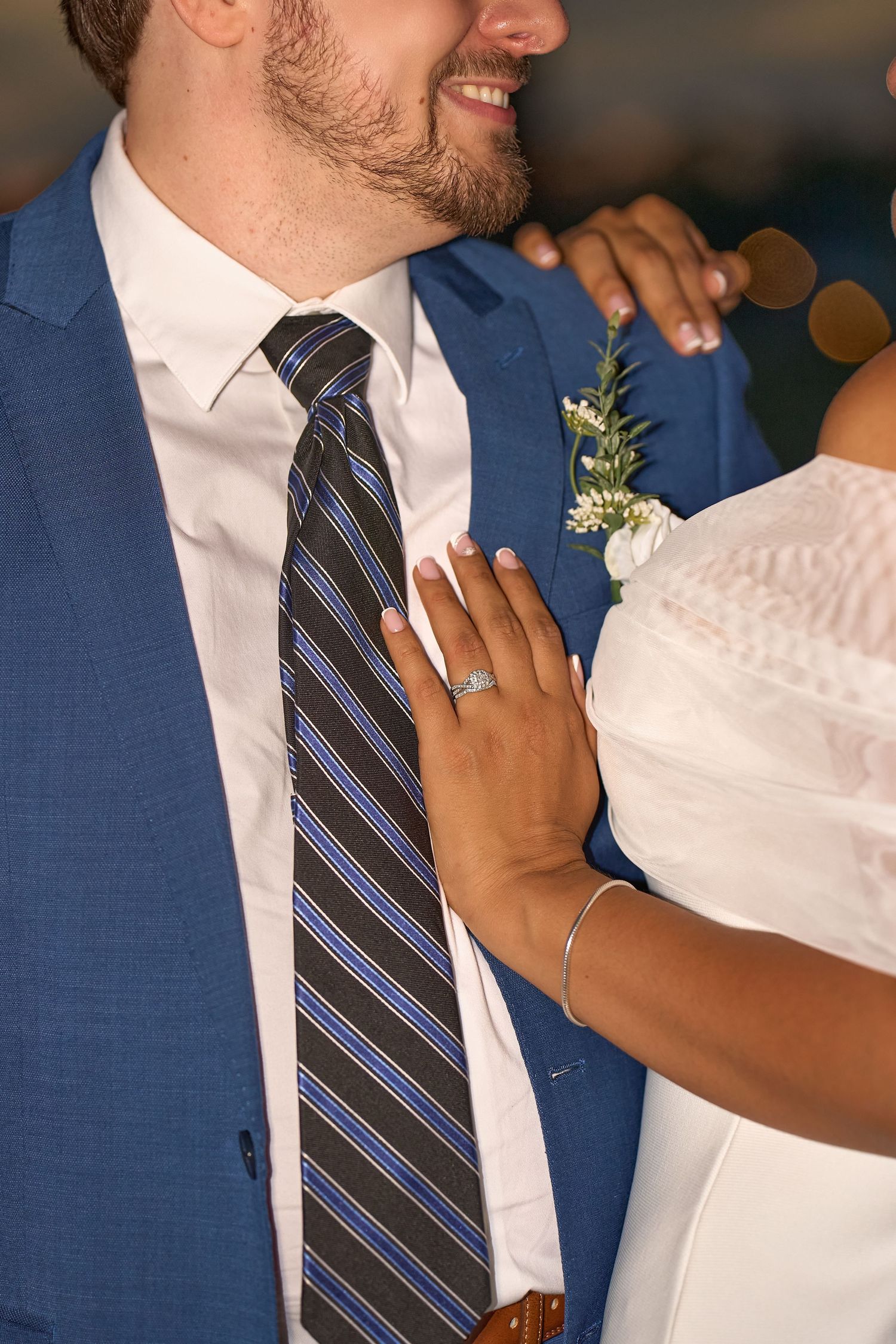 Bride placing hand on groom's chest, showcasing engagement ring, groom in blue suit with striped tie and boutonniere.